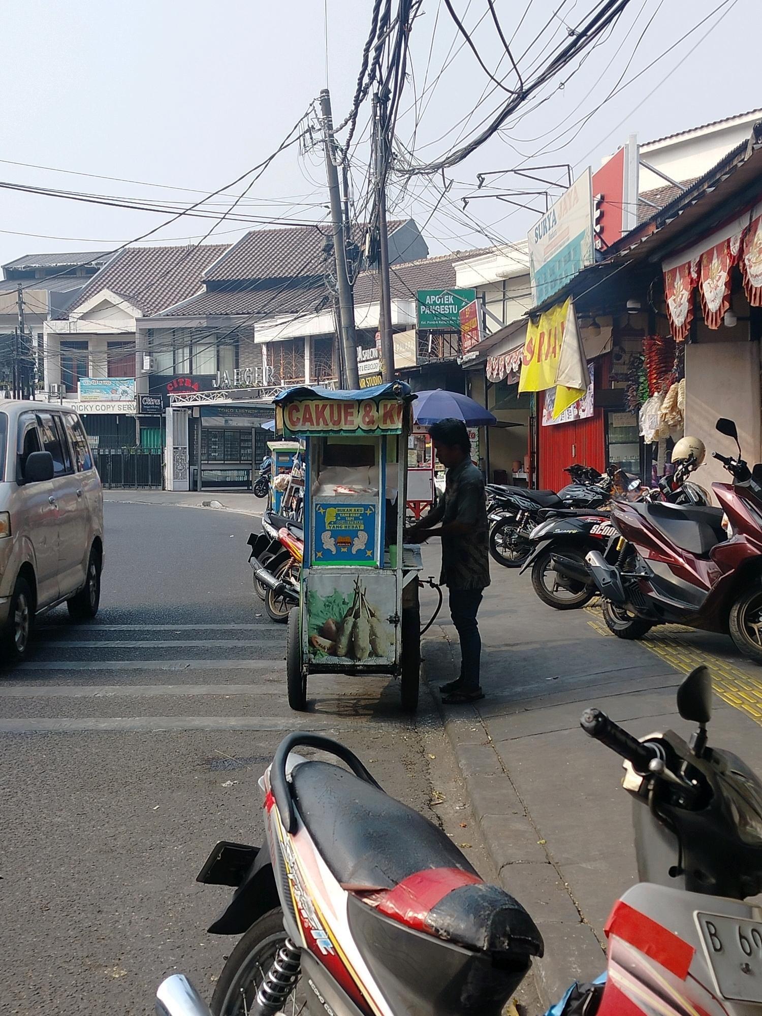 The image depicts a street scene in East Jakarta, Indonesia. A man is standing by a food cart selling "CAKUE & K.*." The cart is green and white with colorful decals. Behind the cart, several motorcycles are parked. To the left of the food cart, a silver van is parked on the road, which has a pedestrian crossing. In the background, there are buildings with storefronts and signage, including "SURYA JAYA" and "APOTEK PANGESTU." Overhead, numerous electrical wires crisscross the sky, strung between utility poles. The environment appears to be urban, with a mix of commercial establishments and residential buildings. The lighting suggests it is daytime.