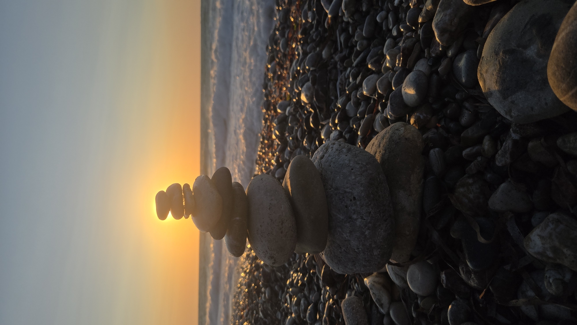A cairn of smooth, grey stones is stacked on a pebble beach during sunset in Soroni, Greece. The sun, a bright orange orb, is visible through the top stones of the cairn, casting a warm glow. The sea is in the background with gentle waves breaking on the shore, and the sky is a soft gradient of orange and blue. The pebbles on the beach are varied in size and color, with some reflecting the light of the setting sun. There are no people or animals visible in the scene.