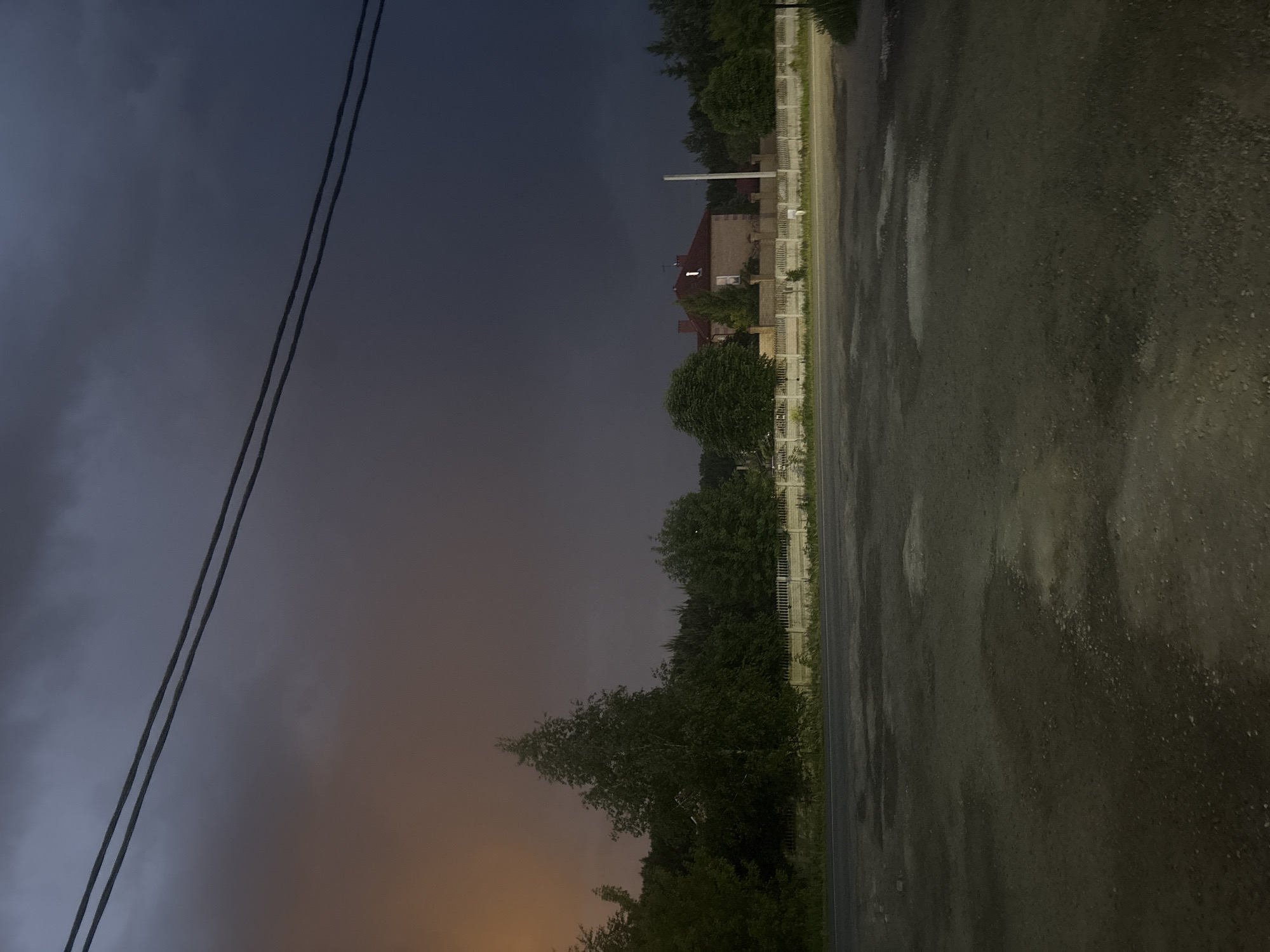 The image, rotated 90 degrees clockwise from its original orientation, portrays a dark, overcast outdoor scene, likely at dusk or under stormy weather conditions. The sky is dominated by heavy, dark clouds, with a discernible orange-yellow hue illuminating the lower left horizon, suggesting either a late sunset or light pollution.
In the foreground, a wet, unpaved road, composed of dirt and gravel, shows numerous puddles and reflective areas, indicating recent rain. Parallel to the road on the left is a long, light-colored fence, appearing to be a white picket style. Behind the fence, dense green trees and bushes line the landscape. Partially visible through the trees is a house featuring a reddish-brown pitched roof and light-colored walls. A tall, slender white pole stands prominently near the house, extending above the treeline.
Two dark utility lines or power cables stretch diagonally across the upper left portion of the dark sky. No people or visible text are present in the image. The scene depicts a quiet, potentially rural or suburban environment in Ореховка, Russia.