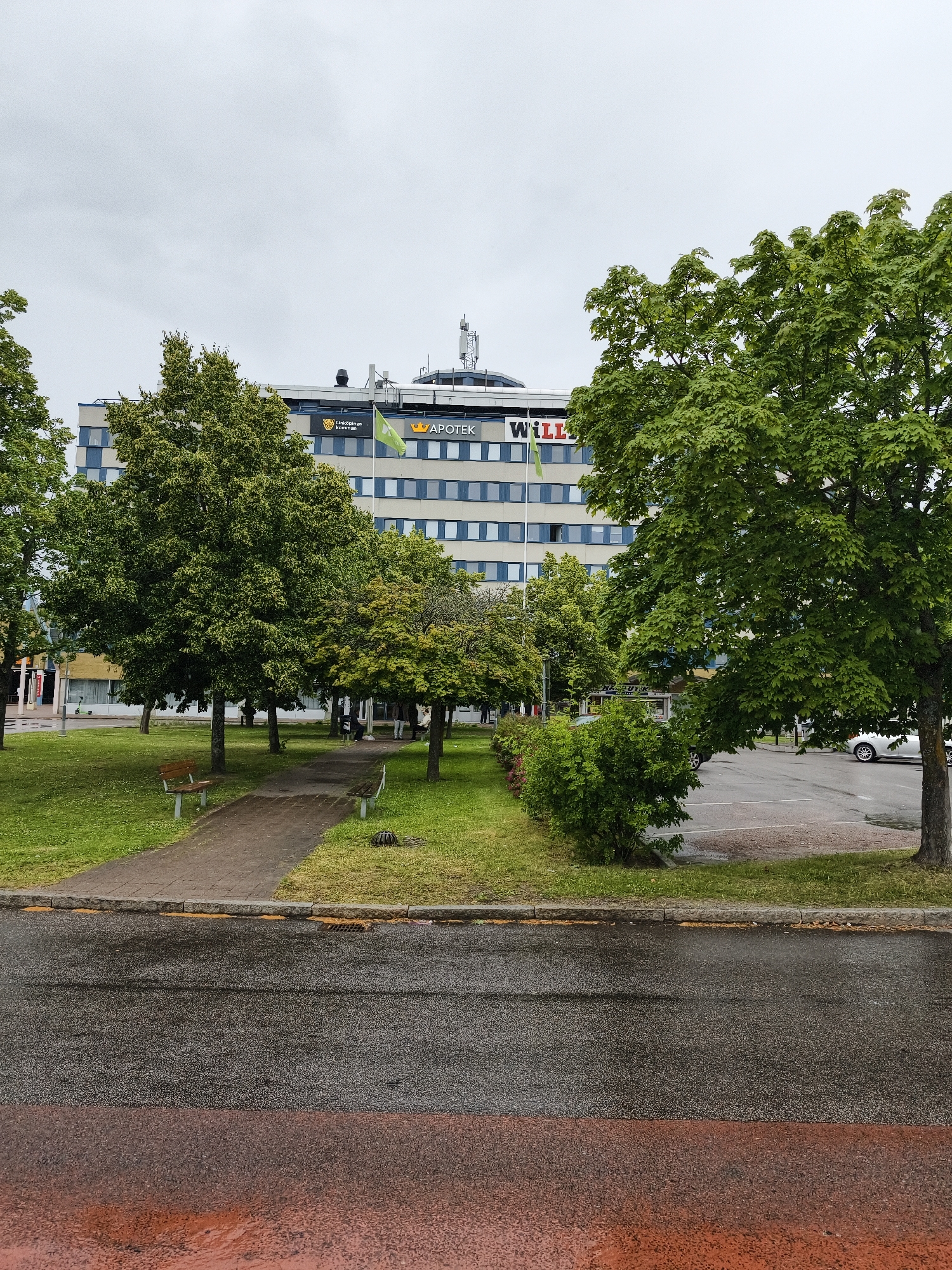 This image depicts an outdoor scene in Linköping, Sweden, on a cloudy day. The foreground is dominated by a wet asphalt road, with a reddish-brown paved area to the very bottom. Beyond the road is a grassy area with a paved walkway and two park benches. Several trees with lush green foliage are scattered throughout the scene, with one particularly large tree on the right side of the frame.
In the background, a multi-story building with a light-colored facade and numerous windows is visible. The building has signs on it, including "Linköpings kommun" and "APOTEK" (which translates to "pharmacy" in Swedish). Another sign reads "WILLI". The overcast sky suggests it might be overcast or raining, contributing to the wet appearance of the road. The scene appears to be a public area, possibly a park or urban green space adjacent to a commercial or municipal building. There are a few people visible in the distance near the building, but their activities are not discernible. The overall mood is subdued, likely due to the weather.