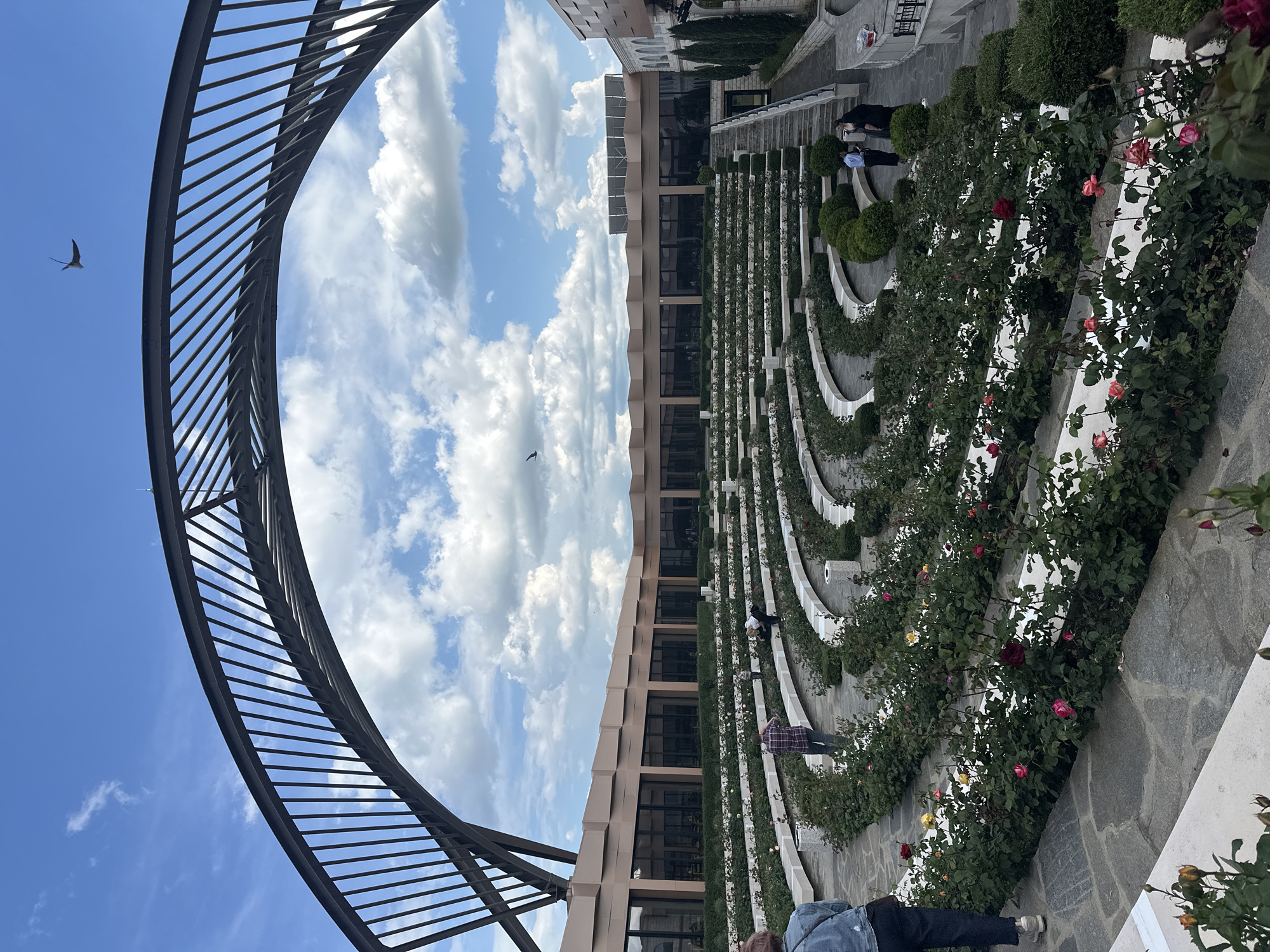 The image shows a garden in Skobelevo, Bulgaria. The garden features rows of rose bushes planted in curved terraces. Several people are present, including individuals walking or standing along the pathways. A large, arched metal structure is visible in the foreground, framing the sky with scattered clouds. A bird is seen flying in the sky. A building with multiple windows stands behind the garden. The ground is paved with stone tiles.
