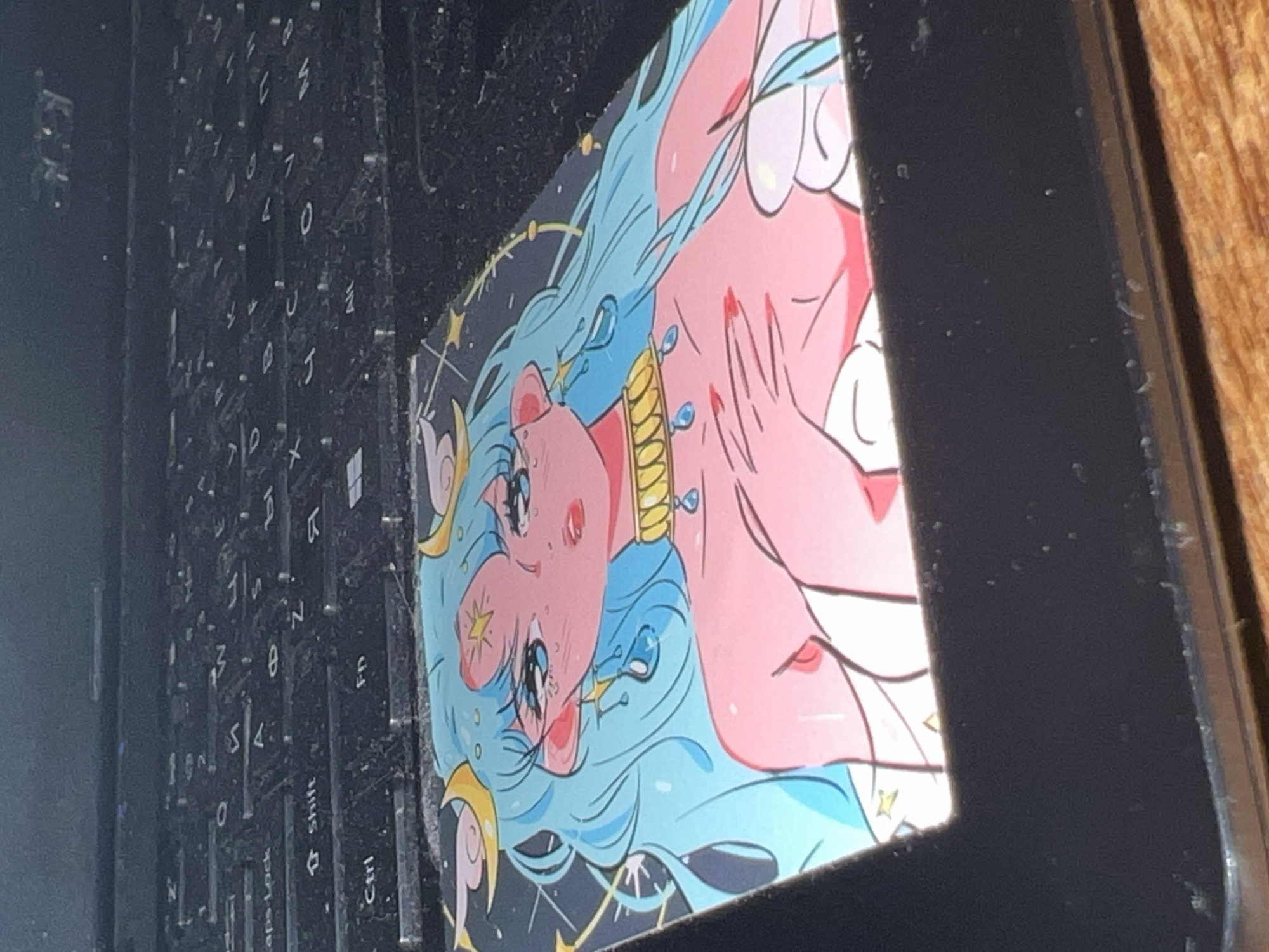 The image shows a black laptop with a sticker on the top right corner. The sticker features an anime-style character with blue hair, fair skin, and jewelry. The background of the sticker is black with yellow stars and crescent moons. The laptop keyboard is visible on the left side of the image. The surface beneath the laptop appears to be made of wood. No location information about Votkinsk, Russia is provided or discernible from the image.