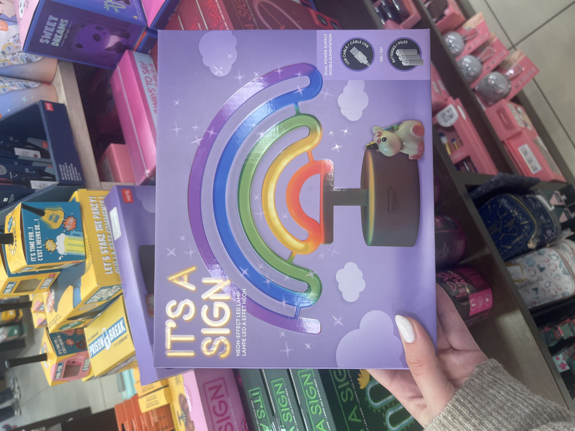 The image shows a hand holding a boxed "It's A Sign" Neon-Effect LED Lamp with a rainbow design and a unicorn figure attached. The packaging indicates it uses either USB cable or batteries for power. The scene is likely inside a retail store, possibly in Varna, Bulgaria, due to the presence of other boxed items and shelves in the background, suggestive of a store environment. However, without additional visual or textual clues pinpointing the specific location in Varna, Bulgaria, one cannot be certain where in the city this picture was taken. Other products around it on the shelves include other items such as "Sweet Dreams" and "Ways to Say" game boxes. A "Prison Break" game box is also visible in the background.