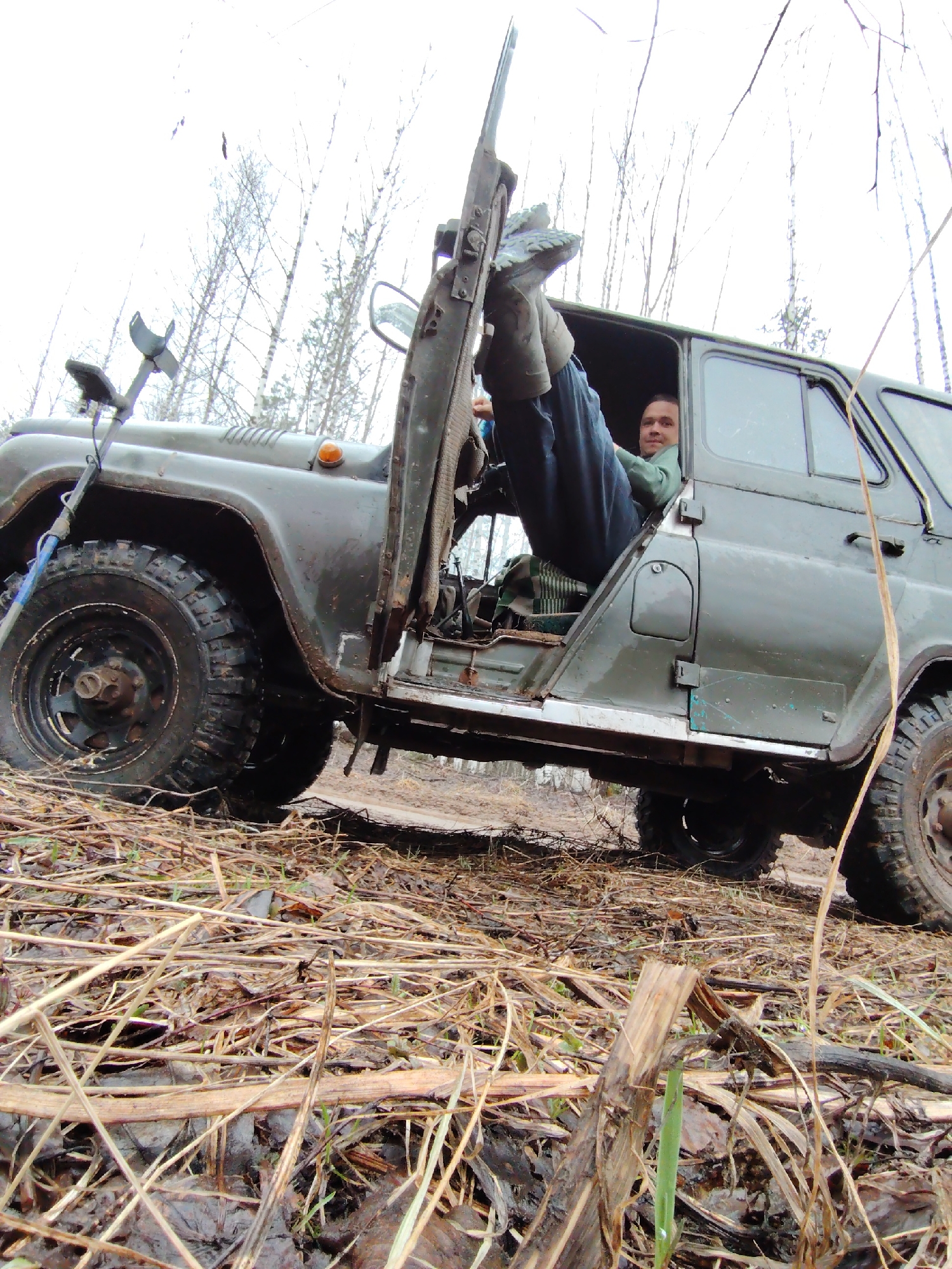 The image depicts a man sitting inside an olive green UAZ-469 vehicle with the door open, parked on uneven terrain covered with dead grass and small plants, near Markovskii, Russia. He has his legs up, with his feet resting against the open door. The man appears to be looking at the camera. A metal detector is attached to the front of the vehicle. The background shows a forest with bare trees under an overcast sky. The ground appears muddy.
