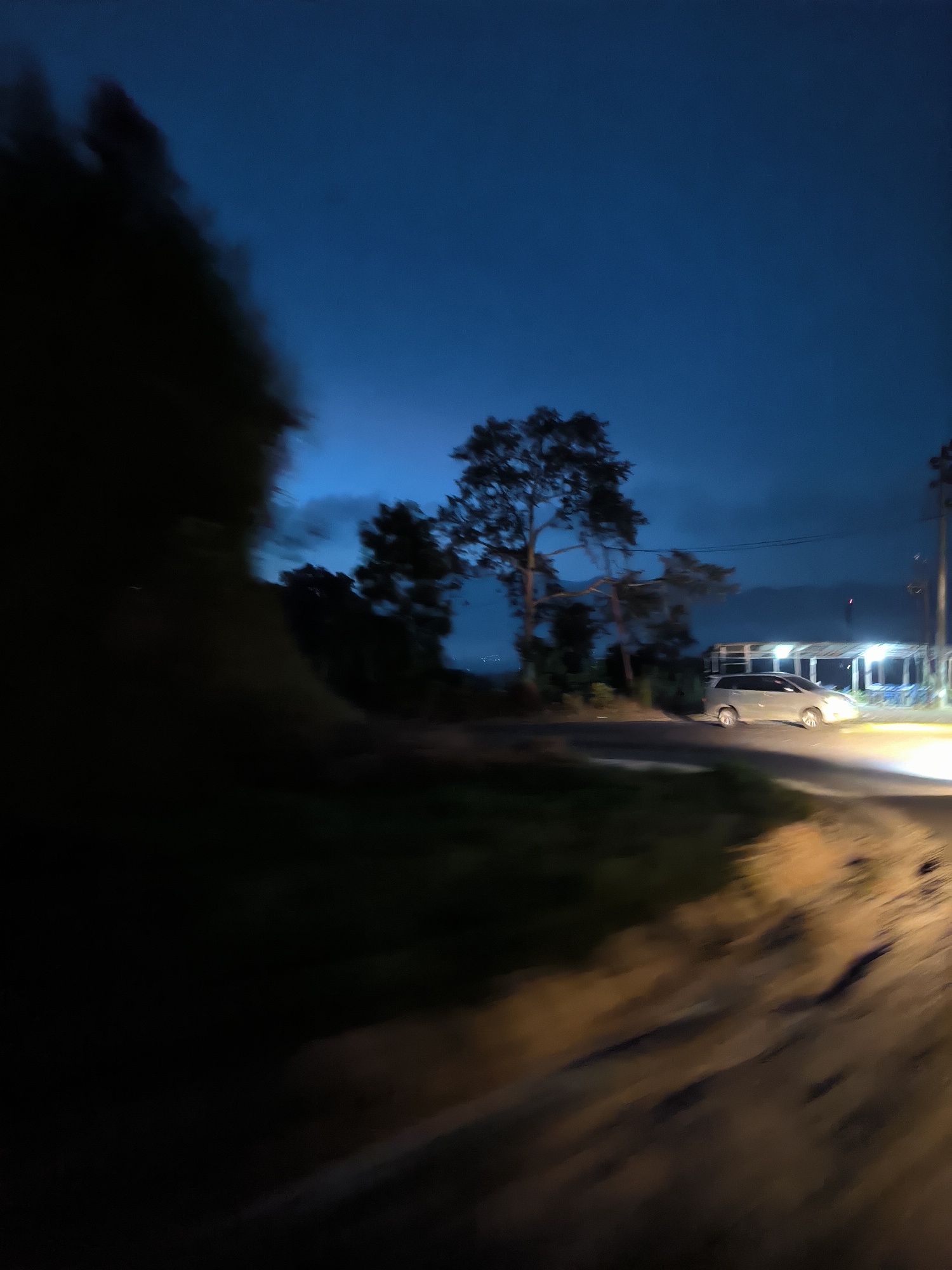 The image captures a twilight scene on a road in Mutiarabaru, Indonesia. The sky is a deep blue, transitioning to a lighter blue near the horizon, suggesting the end of the day. On the left side of the frame, a dark, blurry mass, possibly foliage or trees, dominates the foreground, creating a sense of depth and motion blur. To the right of this mass, a lighter-colored minivan is parked or moving slowly on the road. Beyond the minivan, there is a building with bright artificial lights illuminating an open area, possibly a roadside stall or rest stop. The road surface appears to be unpaved or dirt, with visible tire tracks and debris, and it curves towards the right side of the image. Tall trees with sparse foliage are scattered throughout the background, silhouetted against the sky. A utility pole with wires is visible on the far right. The overall impression is a dimly lit, rural or semi-rural setting.
