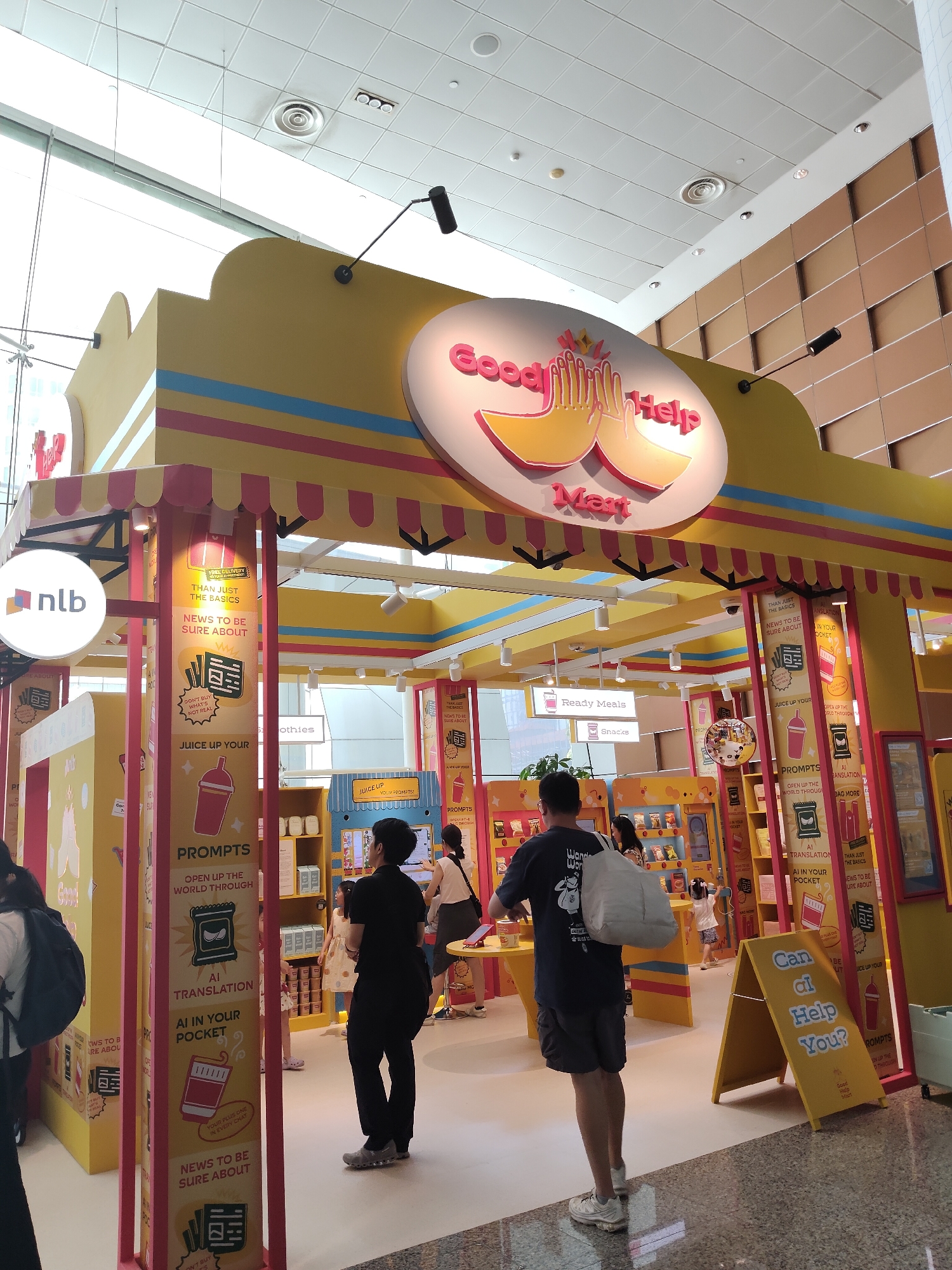 The image shows an indoor marketplace or exhibition booth with a vibrant yellow and pink color scheme, branded "Good Help Mart." The booth features displays with graphics and text related to "Juice Up Your Prompts," "AI Translation," and "News to be Sure About." Several individuals are visible, including a person in a black t-shirt and shorts carrying a white bag, another person in a black t-shirt and dark pants, and a child in a patterned dress. The background includes shelves stocked with items and signage. A yellow A-frame sign in the foreground reads "Can AI Help You?". The environment appears to be a modern, well-lit indoor space, possibly a mall or convention center, with a tiled floor and a visible ceiling with lights and air vents. The "nlb" logo, indicating the National Library Board of Singapore, is visible on a circular sign on the left side of the booth. Based on the presence of the NLB logo and the general ambiance, this scene is likely located in Singapore.