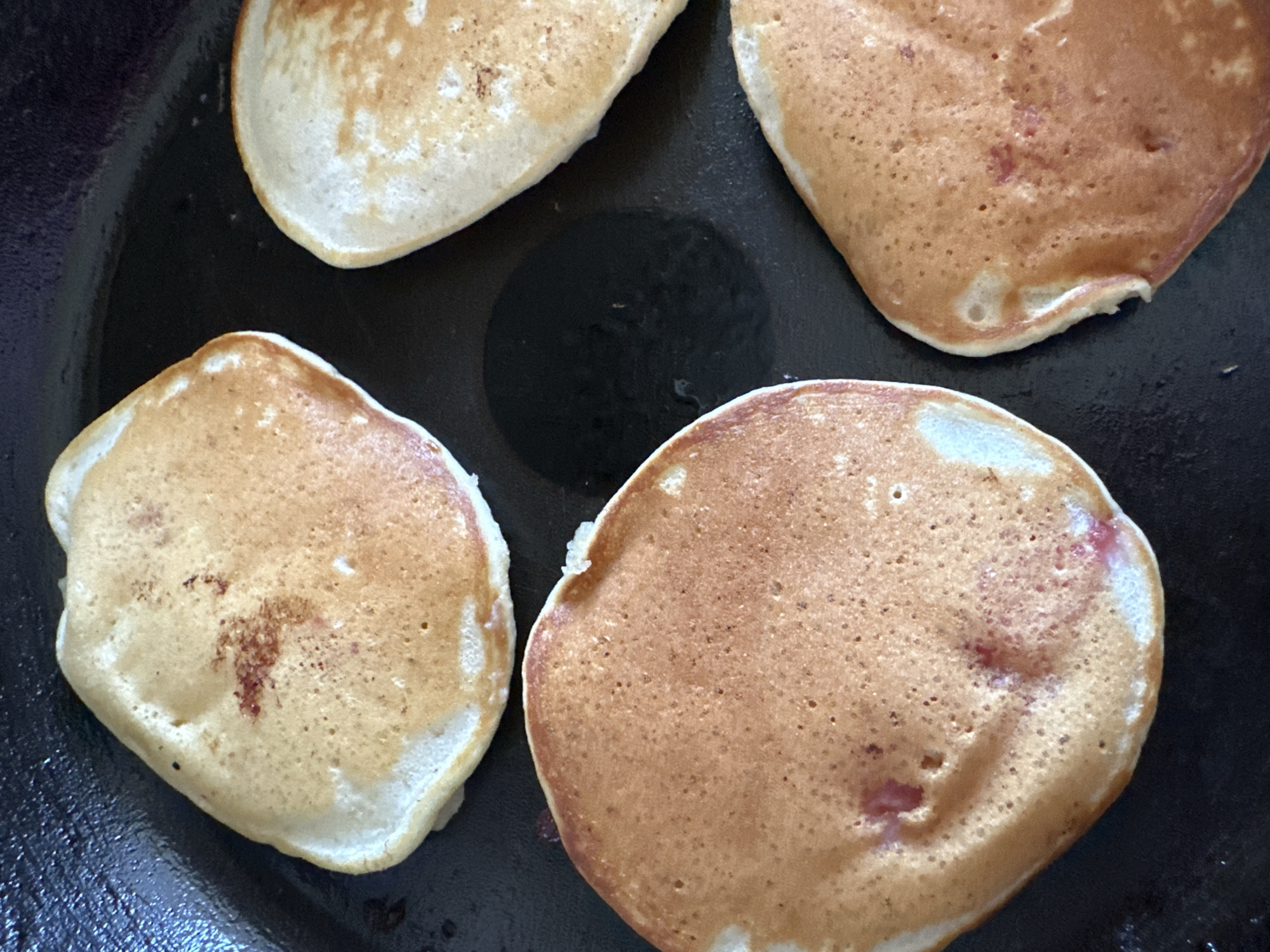 Four golden-brown pancakes are cooking in a dark skillet. The pancakes are circular and have a slightly uneven texture with small bubbles on the surface, indicating they are in the process of being cooked. Some of the pancakes have reddish specks, possibly from fruit mixed into the batter. The skillet appears to be black and has a slightly oily surface. The lighting suggests an indoor setting, possibly a kitchen, and the image is a close-up shot, focusing solely on the pancakes and the skillet. There is no visible text or any contextual information about the time of day, weather, or emotions. The scene depicts the activity of cooking pancakes.