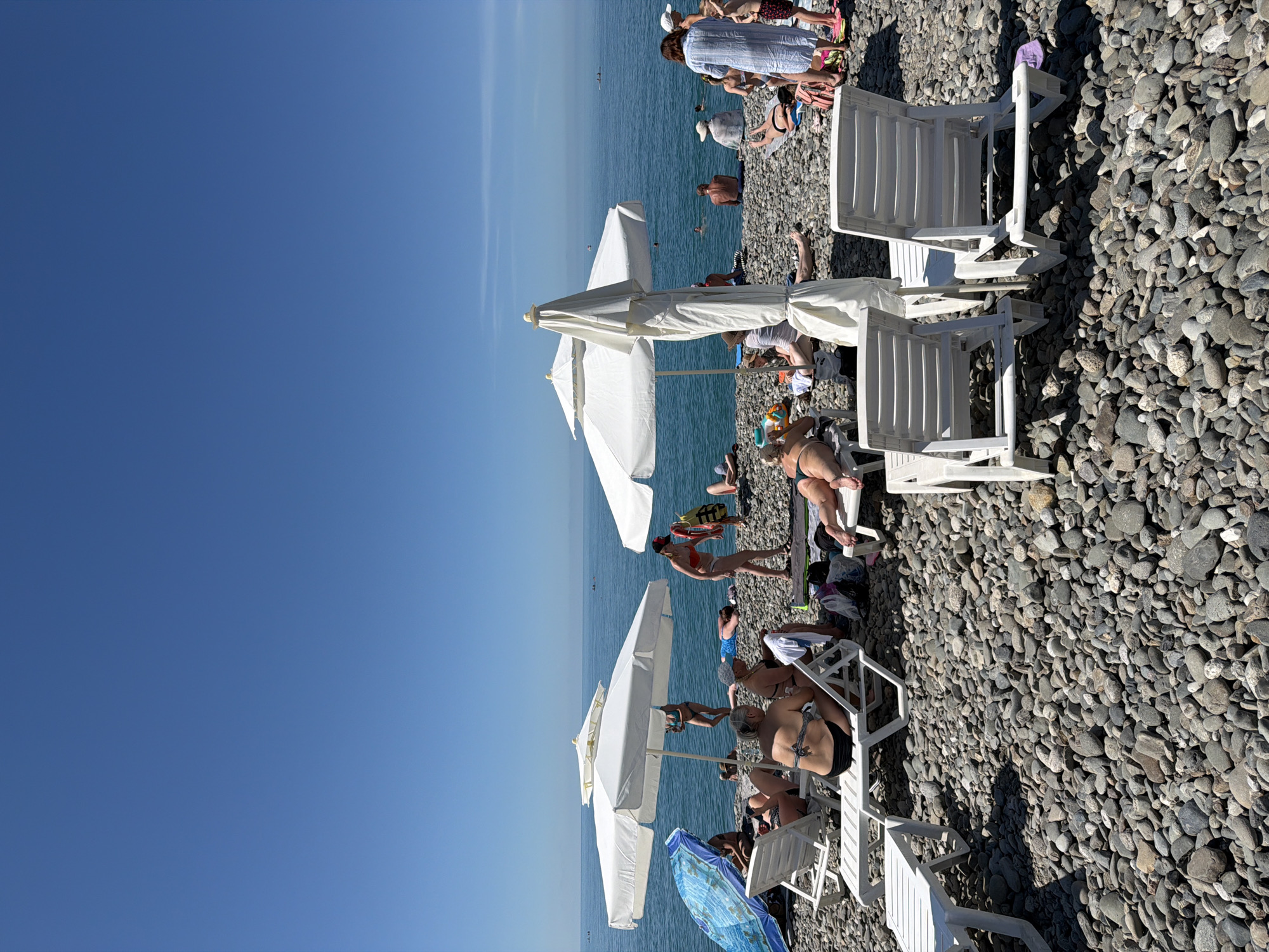 This is a medium shot, taken on a sunny day, of a crowded pebble beach in Sochi, Russia, on the Black Sea. The sky is clear and blue. The water is calm and a deep blue. In the foreground, two white plastic lounge chairs are empty, facing away from the viewer. Behind them, another white lounge chair is occupied by a person wearing a black bikini. To the left, a blue and white striped beach umbrella is open. Further back, several other white beach umbrellas are open, providing shade for beachgoers. Many people are sunbathing or relaxing on lounge chairs or directly on the pebbles. Some people are in the water. To the right, a woman in a blue and white striped dress is standing and looking towards the water. A life vest is visible on her arm. Several bags and towels are scattered on the beach. The overall scene depicts a typical summer day at a popular tourist destination.