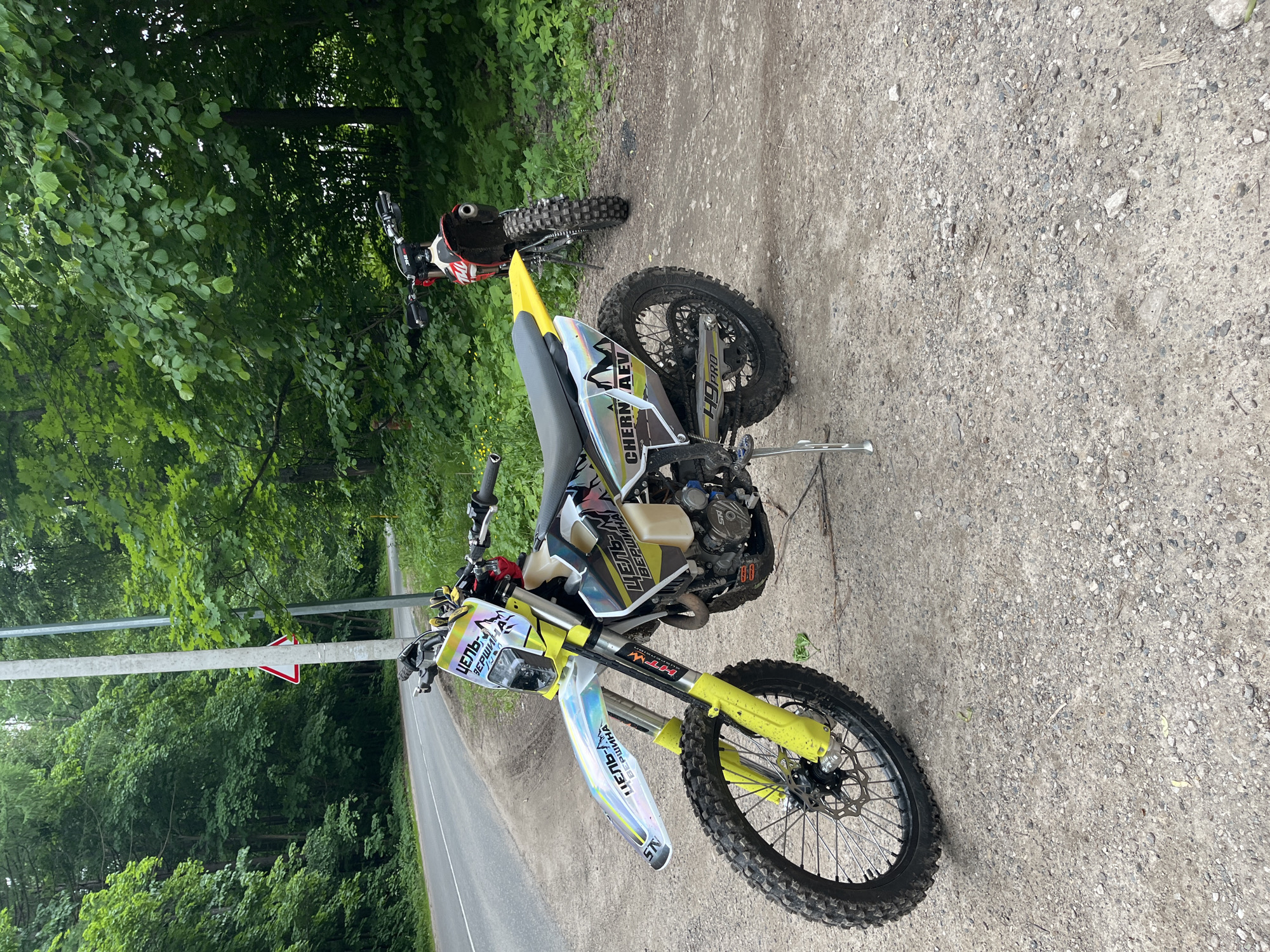 This image displays two dirt bikes parked on a gravel and dirt shoulder beside an asphalt road in Ореховка, Russia. The foreground motorcycle is a yellow and white enduro bike, prominently featuring numerous stickers including "ЦЕЛЬ ВЕРШИНА" (Target Summit) on the front fender and headlight cowl, "CHERN AEV" on the rear side panel, and "H9 PRO" on the swingarm. It is equipped with knobby tires, yellow front suspension forks, disc brakes, and a deployed kickstand, exhibiting accumulated dirt consistent with off-road use. A second, red and white dirt bike is partially visible further in the background, also parked on the gravel shoulder. The asphalt road, marked with a solid white edge line, curves into the distance and is bordered on both sides by a dense, verdant forest comprised of mature deciduous trees and thick undergrowth. A white triangular road sign with a red border, likely indicating a hazard, is mounted on a light-colored utility pole positioned behind the front motorcycle. No people are present in the scene.