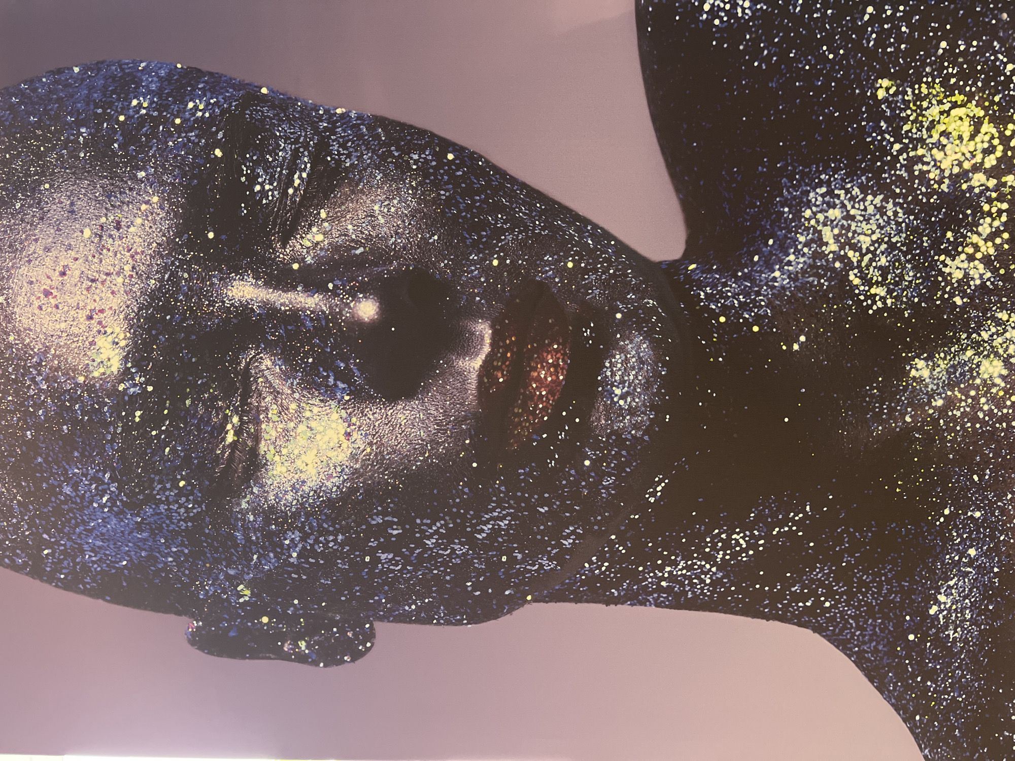 The image is a close-up, full-frame portrait of a person covered in glitter. The person's face, neck, and shoulders are visible. The glitter is predominantly dark blue and purple, with accents of yellow and gold, creating a cosmic or starry effect. The person's eyes are closed, and their lips are highlighted with a shimmering, reddish-gold glitter. The background is a soft, gradient of pale pink and purple. There are no discernible landmarks or contextual clues to suggest a specific location in Vitsebsk, Belarus.