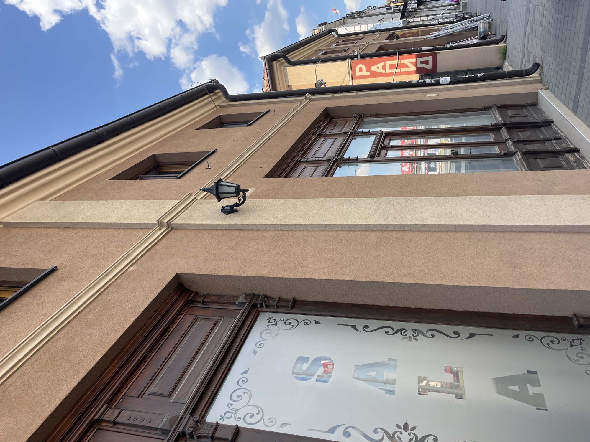 This is a medium-angle, eye-level, outdoor shot of a row of buildings on a sunny day in Čačak, Serbia, taken from a sidewalk. The foreground features the side of a building with a light brown stucco exterior. The building has a large, dark wooden door with ornate carvings and frosted glass. The glass has a decorative border and the letters "S" and "A" are visible, possibly part of a larger word or logo. To the right, the building continues with a darker brown stucco and a tall, narrow window with dark wooden frames. Further down the street, other buildings are visible, one with a red sign that has white Cyrillic lettering, reading "РАДИО" (RADIO). The sidewalk is paved with grey rectangular bricks. In the background, there are more buildings, a bicycle, and a Serbian flag flying from a pole. The sky is blue with scattered white clouds.