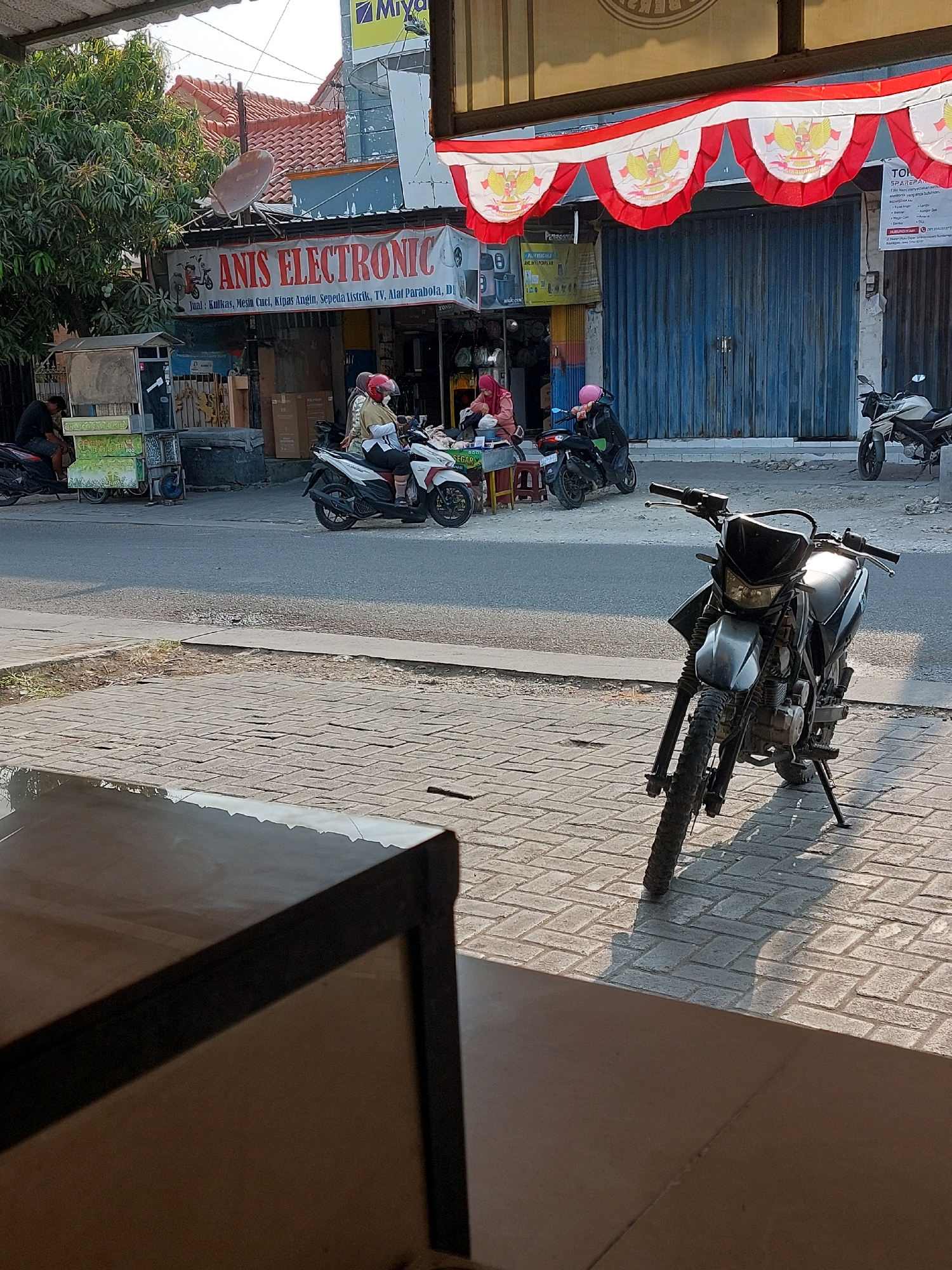 The image depicts a street scene in Sumuragung, Indonesia. In the foreground, a black dirt bike is parked on a brick sidewalk. The bike is angled slightly towards the viewer, with its front tire prominent. Behind the bike, a road is visible, with several motorcycles and people. A white motorcycle with two individuals, one wearing a yellow shirt and helmet, and another wearing a pink helmet, is stopped on the road. Another person in a pink helmet is sitting on a black motorcycle. To the left, a person in a black shirt is seated on a motorcycle.
The background features shops with awnings. One awning displays "ANIS ELECTRONIC" with text underneath detailing products like refrigerators, washing machines, fans, TVs, and parabolic antennas. Another shop has a blue metal roll-up door. A satellite dish is mounted above the "ANIS ELECTRONIC" awning. Red and white pennant banners, adorned with Indonesian national emblems, are strung across the front of the shops. Trees and buildings are also visible in the distance. The scene appears to be illuminated by the late afternoon sun, casting shadows on the brickwork.