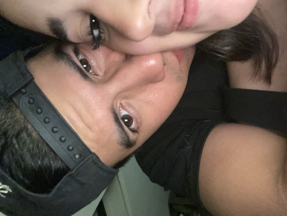 The image displays a close-up of two individuals in an intimate pose within an unidentifiable indoor setting. The scene is dimly lit.
The primary subject, a male, occupies the left and central portion of the frame. He is wearing a black baseball cap backward, revealing the black plastic snapback adjuster. His hair is dark and short. He has dark brown eyes, a prominent nose, and appears to be looking directly at the camera. He is wearing a black short-sleeved shirt, on which several light-colored fibers or lint are visible near his right shoulder and upper arm. His right arm and shoulder are prominent in the lower left.
The second subject, a female, is positioned to the right of the male, with only the right side of her face visible. Her right eye has long, dark eyelashes, and she possesses a slight smile. Her dark brown hair cascades along her right cheek and neck. The male's face is pressed against her right cheek/nose area, indicating close physical proximity. Both individuals exhibit features consistent with Hispanic ethnicity.
The background is dark and out of focus, providing no discernible details about the specific room or surroundings. A faint, lighter-toned object or surface is vaguely visible in the upper left background.
The image provides no visual information to determine its location within the city of Coatzacoalcos, Mexico.