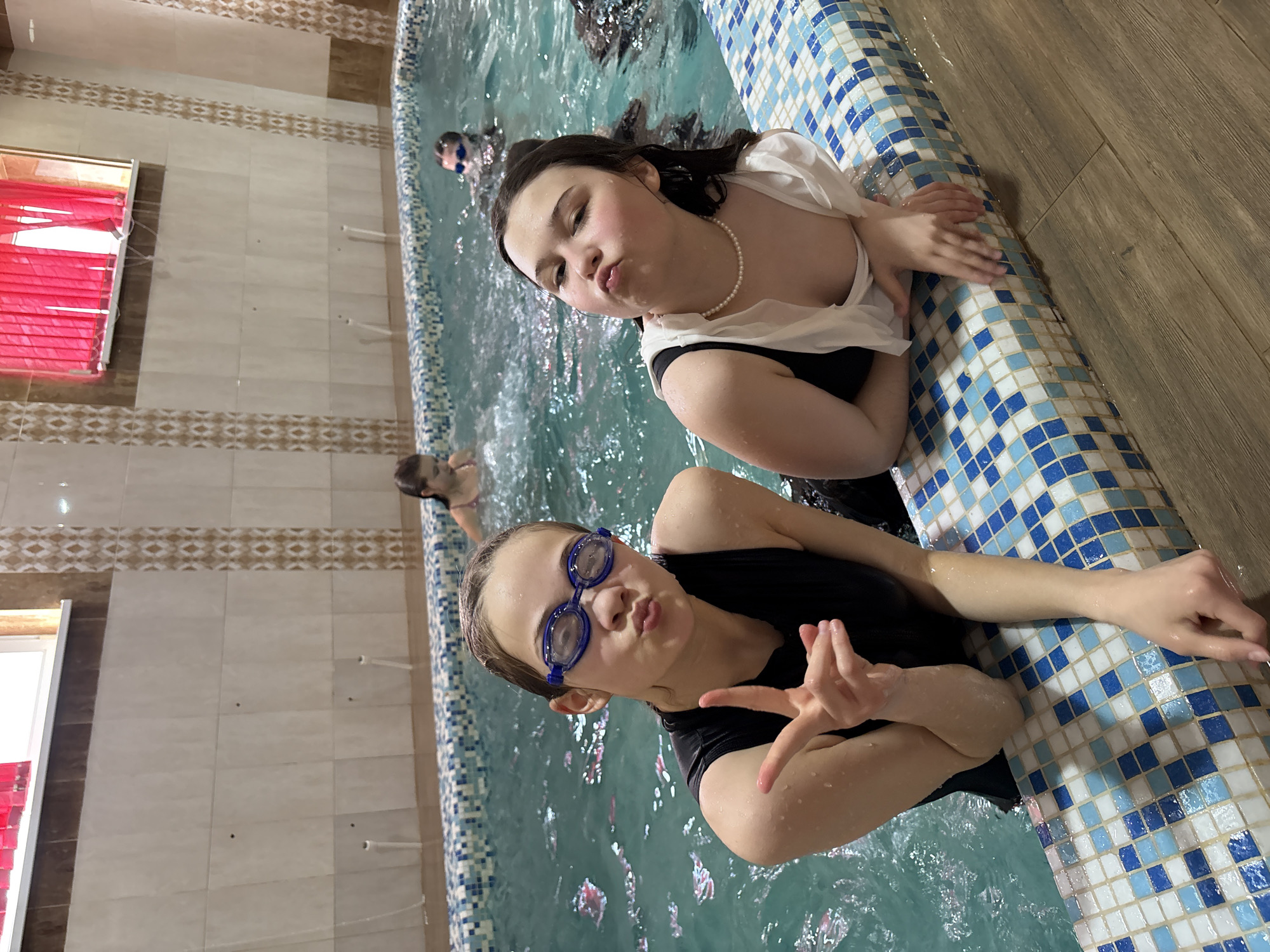 An indoor swimming pool scene in Chaikovsky, Russia, features two young girls posing playfully for the camera. The image is taken from a slightly elevated, diagonal perspective, looking down into the pool.
In the foreground and midground, two girls are submerged in the clear, light blue water of the pool. The girl in the foreground, closer to the camera, wears blue swimming goggles on her head and a dark swimsuit or top. She has wet, light brown hair, is making a "peace sign" or similar hand gesture with her right hand, and is puckering her lips in a playful expression. Her left arm is resting on the mosaic-tiled edge of the pool.
The second girl, slightly further back, has dark wet hair and is wearing a white blouse or cover-up over a dark swimsuit or top, along with a white pearl necklace. She is also puckering her lips in a similar playful manner and has her arms resting on the pool's edge. Both girls appear cheerful and engaged with the camera.
Other individuals are partially visible in the water in the background, suggesting a group activity. The pool edge is adorned with blue and white mosaic tiles arranged in a checkered pattern. Beyond the pool, to the left, is a wall covered in light beige square tiles, featuring a horizontal decorative border of brown and light-patterned tiles. A window with bright red horizontal blinds is visible on this wall. The area immediately surrounding the pool to the right has a light brown wooden plank floor. No visible text is present in the image.