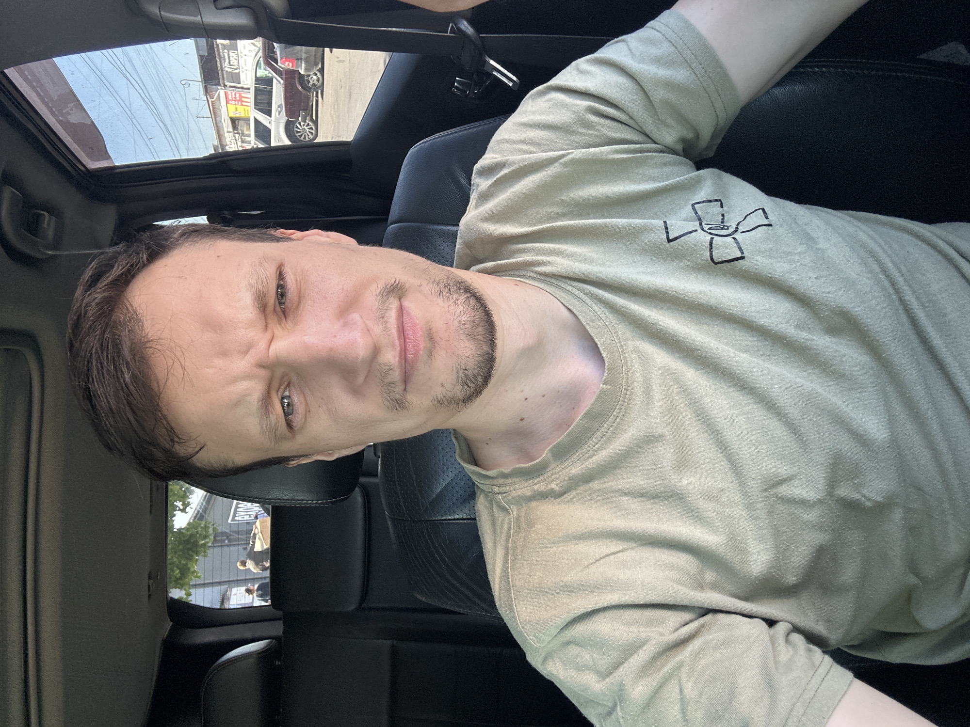 A man with light skin, dark brown hair, and a goatee is sitting in the driver's seat of a car, taking a selfie. He is wearing a light brown t-shirt with a black symbol on the left breast. The interior of the car is visible, including black leather seats, a sunroof, and the car door. Outside the car, through the windows, a street scene is visible with buildings, parked cars, and power lines. The presence of a cross symbol on the man's shirt may suggest a connection to Ukraine, as crosses are a common symbol in Ukrainian culture and religion. The car appears to be parked on a street in Kamianets-Podilskyi, Ukraine, given the context provided.