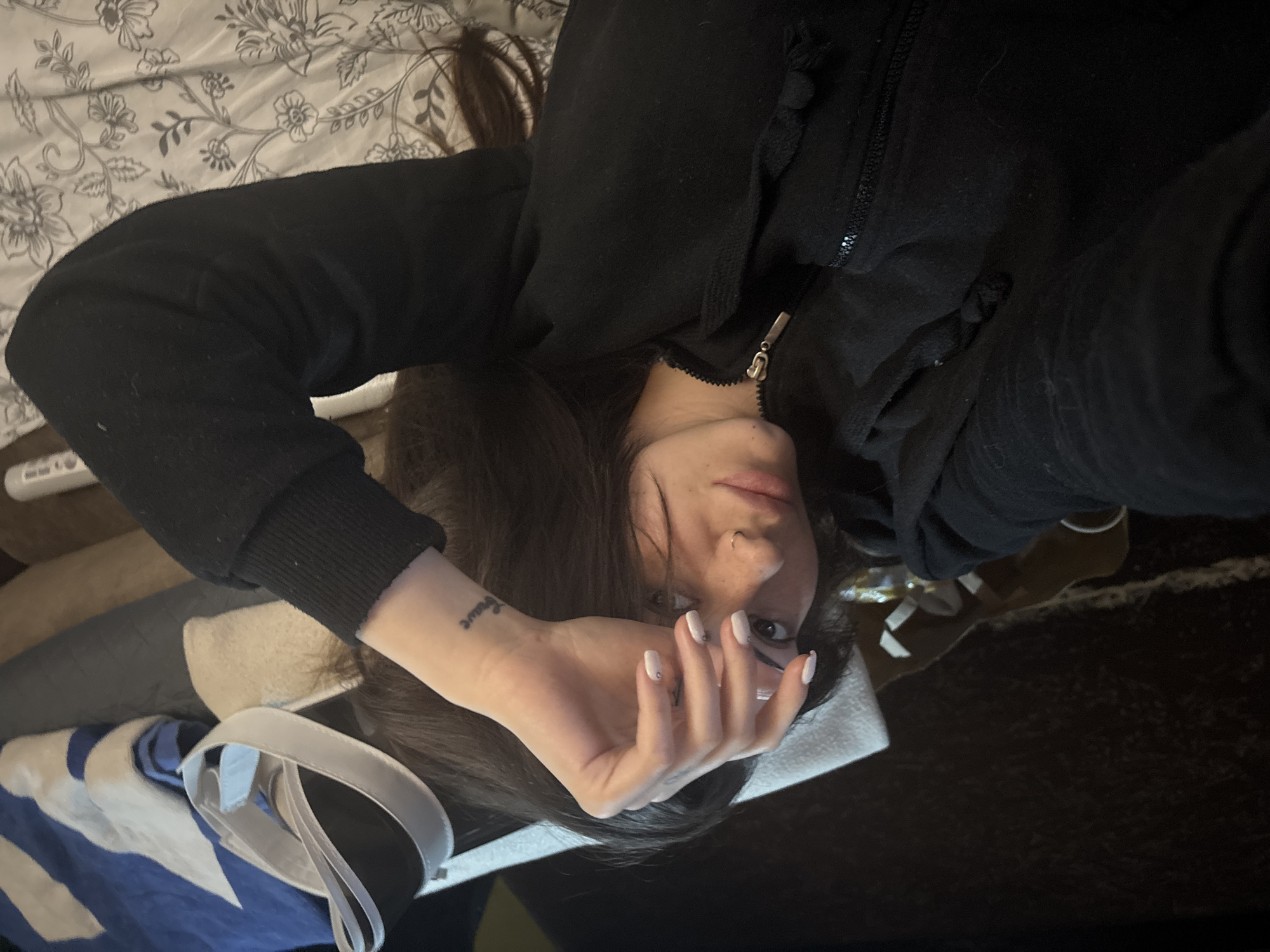 An indoor portrait captures a person, likely female, lying down, appearing to take a selfie in Belopoptsi, Bulgaria. The image is rotated 90 degrees clockwise, showing the person's head to the left. They have long, dark hair and are wearing a black hooded jacket with a zipper and drawstrings. Their left hand, featuring light-colored nail polish with small dots, is raised and partially obscuring the face, with fingers slightly spread. A small tattoo reading "brave" is visible on the inner wrist of this hand. The person has a nose piercing and looks directly at the camera with a neutral gaze. Behind them, a white and grey floral patterned bedding or blanket is visible, suggesting the person is on a bed. Other objects in the background include blue and white striped fabric, a white strap, and indistinct dark surfaces. The lighting is soft and consistent with an indoor setting.