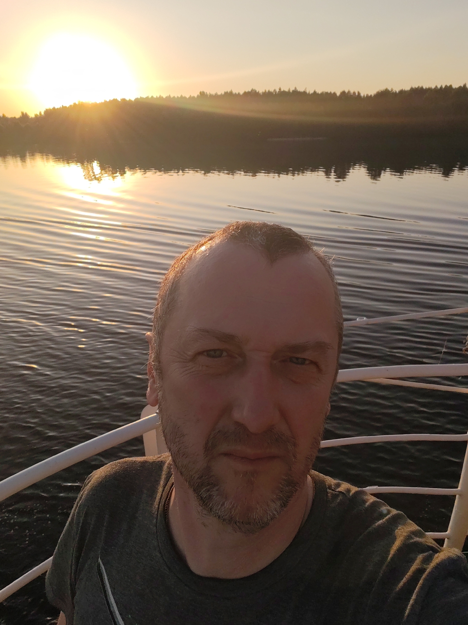 A man is taking a selfie on a boat. The background shows a body of water with a treeline and a bright sun. The man has short, graying hair and a beard, wearing a gray t-shirt. The boat has a white railing that's partially visible. It appears to be a sunset or sunrise over a lake or river. There is no information to support the picture was taken in the city of Vaulino, Russia.