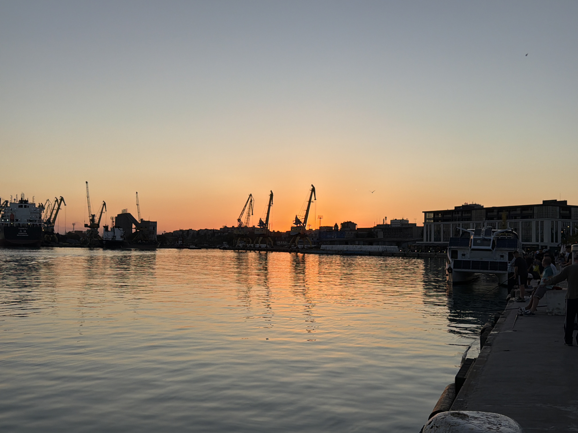 The image shows a harbor scene at sunset in Burgas, Bulgaria. In the foreground is the calm water reflecting the orange and yellow hues of the setting sun. Several cranes and ships are visible along the coastline. People are standing on the dock. A boat is docked nearby. The sky is a gradient of colors transitioning from orange to light blue. The buildings of the city are silhouetted against the sky.