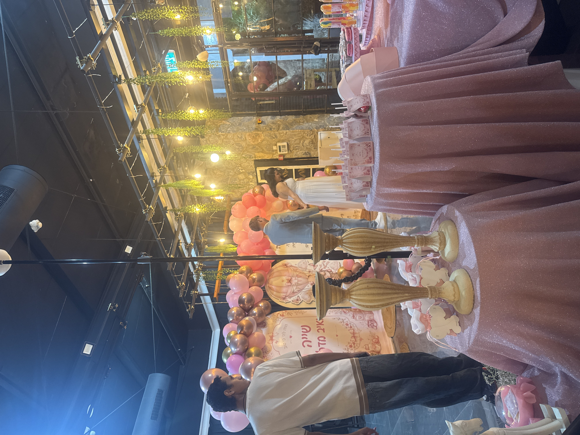 The image shows an indoor event space in Migdal HaEmek, Israel, decorated for a celebration. The primary focus is a dessert table adorned with pink, glittery tablecloths, and various sweets. Among the desserts are macarons, cake pops, and other confections displayed on tiered stands. Two ornate candle holders with beige bases and dark accents are placed on a round table in the foreground. A backdrop featuring balloons in shades of pink and gold, along with a graphic of a hot air balloon and Hebrew text, suggests a baby shower or similar event. Several small pink bags with white ribbons and elephant-shaped decorations are also visible on the table.

In the background, two men and a woman are present. One man, wearing a white t-shirt and jeans, stands facing away from the camera. Another man in a light grey suit is seen from the side. A woman in a white dress is also visible. The environment is lit by overhead lights and hanging decorative lights that create a warm ambiance. The ceiling is black, with exposed industrial elements like ventilation systems and light fixtures. The walls appear to be made of stone, and there is a large window or glass door in the background with reflections of the event. Green hanging plants add a touch of nature to the decor.