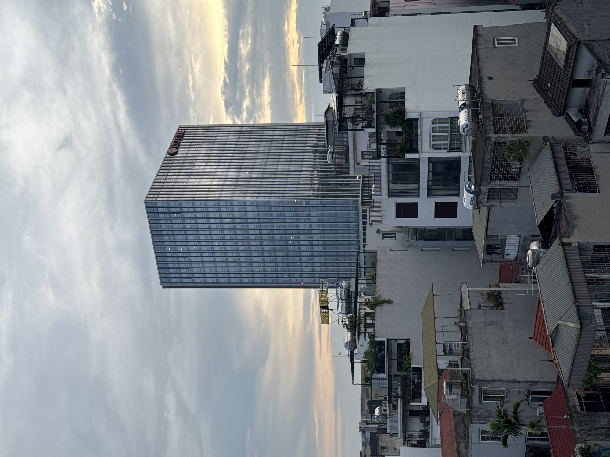 This is an aerial, eye-level shot of a city skyline in Hanoi, Vietnam, at dusk. The main subject is a tall, modern glass-fronted skyscraper with a red "LOTTE" logo near its summit. In front of the skyscraper are several multi-story buildings with flat roofs, many adorned with water tanks and satellite dishes, suggesting a dense urban environment. The buildings range in architectural style from older, weathered structures to more contemporary facades. Several trees, including a palm tree, are visible, along with various roof types, including corrugated metal and tiled. The sky is cloudy, with soft hues of yellow and orange indicating the setting sun breaking through. No people are visible in the image. The scene captures a mix of old and new architecture characteristic of Hanoi.