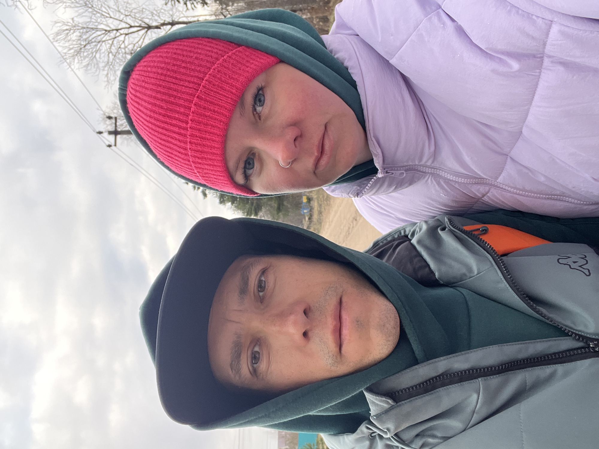 An outdoor portrait features a woman and a man, captured in a selfie-style shot in Chaikovsky, Russia. The image is presented in a portrait orientation, showing the subjects from the chest up against an overcast sky and a natural background.
On the left, a woman with fair skin and blue eyes looks directly at the camera with a neutral expression. She wears a vibrant pink knit beanie over a dark green hooded sweatshirt, and a light lavender puffer jacket. A silver nose ring is visible on her left nostril.
On the right, a man with fair skin and brown eyes also looks towards the camera with a neutral gaze. He is dressed in a dark green hooded sweatshirt and a grey or slate-colored zip-up jacket. A black and white logo, resembling the Kappa brand's 'Omini' symbol, is visible on the upper right side of his jacket. An orange strap or piece of clothing is peeking out from under his unzipped jacket.
The background reveals a hazy, overcast sky with bare tree branches suggesting a colder season (late autumn, winter, or early spring). Overhead power lines and a utility pole are visible behind the woman, and a glimpse of what appears to be a dirt path or road can be seen in the distance between the two individuals. Both subjects are warmly dressed, indicating cool or cold weather. The overall impression is a casual outdoor moment under an overcast daytime sky.