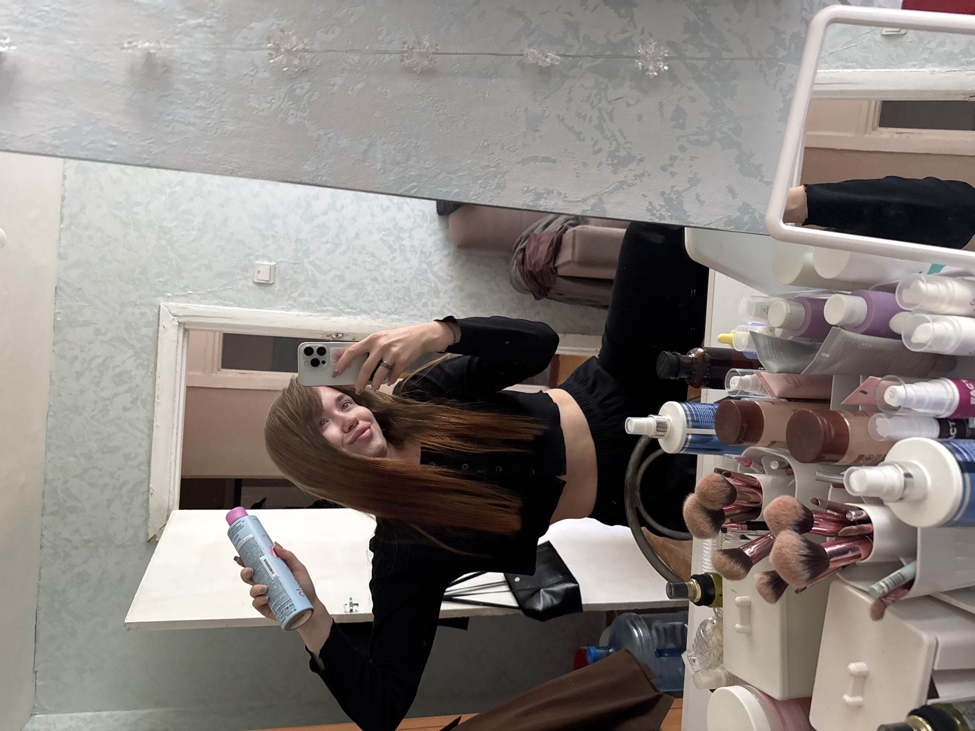 A young woman stands in what appears to be a bathroom in a residential setting, likely in the city of Chaikovsky, Russia. She is taking a selfie in a mirror while holding a blue and pink hair product can in her left hand and her smartphone in her right hand. The woman has long, light brown hair and wears a black cropped top and black pants.
The bathroom has light blue patterned wallpaper, a white doorframe, and a white shelf. The mirror reflects the woman, the bathroom interior, and a glimpse of another room showing what seems to be a bed. A white vanity or storage unit, full of various cosmetic items such as makeup brushes and bottles, is visible to the right of the mirror. A brown jacket is draped over a chair at the bottom of the frame, and partially visible under the vanity are water jugs and containers.