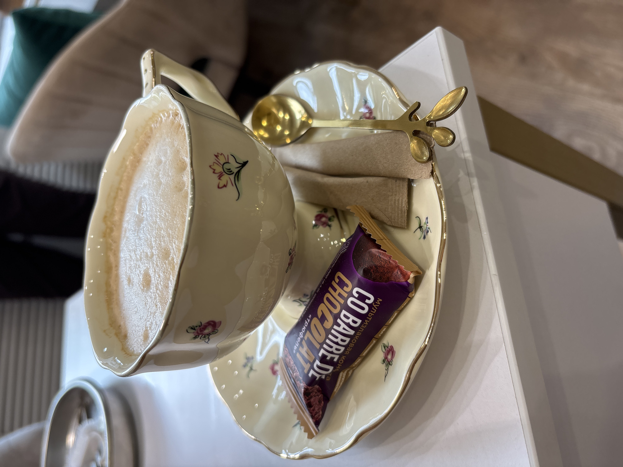 A high-angle, medium shot captures a partially filled teacup and saucer on a white surface. The teacup, made of off-white porcelain, features a floral pattern. The cup contains a light brown beverage topped with foam. The saucer, matching in design to the teacup, holds a small gold spoon, two packets of brown sugar, and a partially unwrapped "Co Barre De Chocolat" chocolate bar. The snack's wrapper is primarily purple with brown and gold accents. The background is out of focus, depicting neutral-toned furniture, a white surface, and possibly a green pillow or cushion. The location cannot be identified as Podosenikha, Russia, from this image alone.