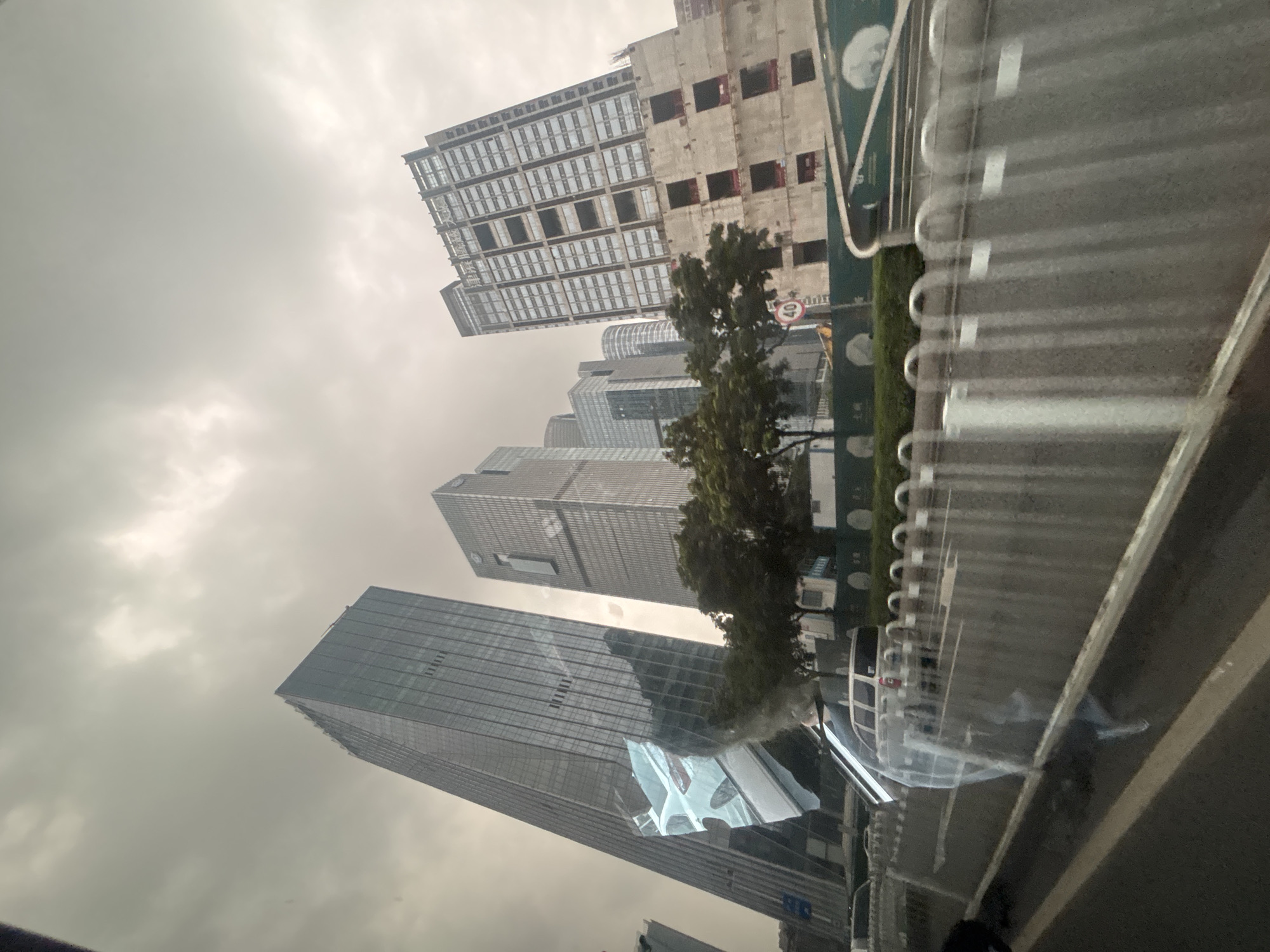 The image depicts a street view in Tianhe District, China, under an overcast sky. In the foreground, a street with a metal fence is visible. Beyond the fence, cars are present, and trees line the roadside. The background is dominated by a cluster of modern skyscrapers. One prominent skyscraper on the right appears to be under construction, with its concrete framework exposed on lower levels. The buildings are characterized by glass facades and a geometric architectural style. Reflections are visible on the glass surfaces of the buildings, including an image of a white car.