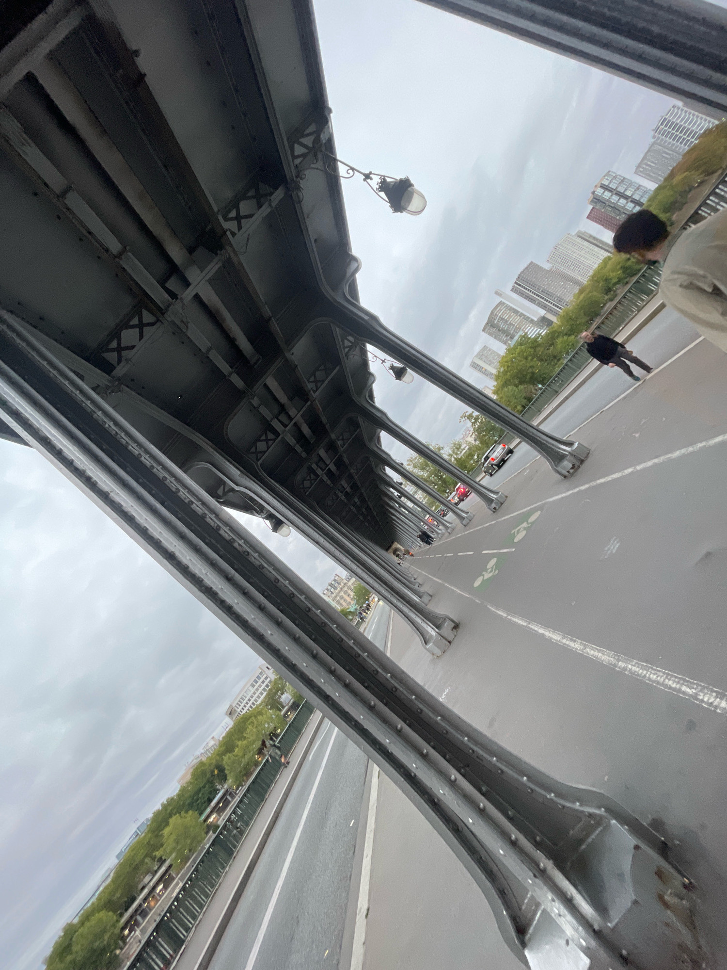 This image was captured on the Pont de Bir-Hakeim bridge in Paris, France. The perspective is from underneath the bridge's structure, looking outwards towards the street and cityscape. The bridge's ironwork, with its repeating arched supports and rivets, dominates the foreground and midground. On the street below, there is a designated bike lane with green markings and bicycle icons. Several people are visible walking on the pedestrian walkway to the right of the bike lane, and a few cars are on the road. In the background, the distinctive modern architecture of buildings can be seen rising above a line of trees, under an overcast sky.