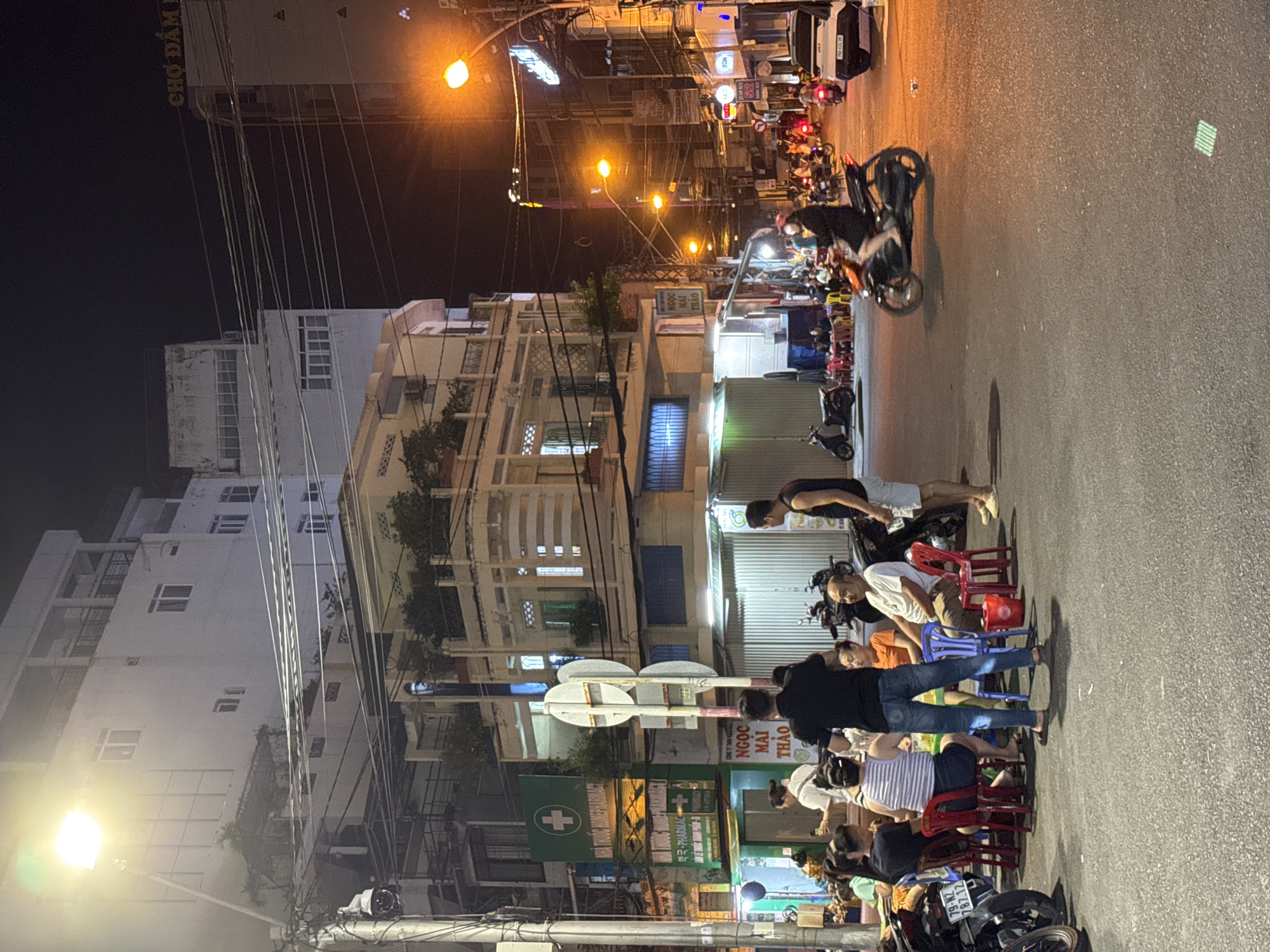 The image depicts a nighttime street scene in Nha Trang, Vietnam, likely near a commercial area. In the foreground, a group of people are seated at small, round tables on the asphalt street, some of them engaged in conversation. They are seated on red and blue chairs. To the left, several motorcycles are parked. A man in a black t-shirt and jeans stands facing away from the camera, while another man in a black tank top and shorts stands to his right.
In the mid-ground, a woman is riding a scooter towards the viewer, her face obscured by darkness and possibly a helmet. More scooters and a white car are visible further down the street, along with multiple streetlights illuminating the scene. The buildings in the background are multi-story structures, some appearing to be apartments or shops, with various signs and lights. One prominent sign displays "NHÀ THUỐC HOMEPHARMACY" in green.
The street is paved with asphalt and appears to be well-trafficked. The atmosphere is characteristic of a bustling urban environment at night, with a mix of residential and commercial activity.
