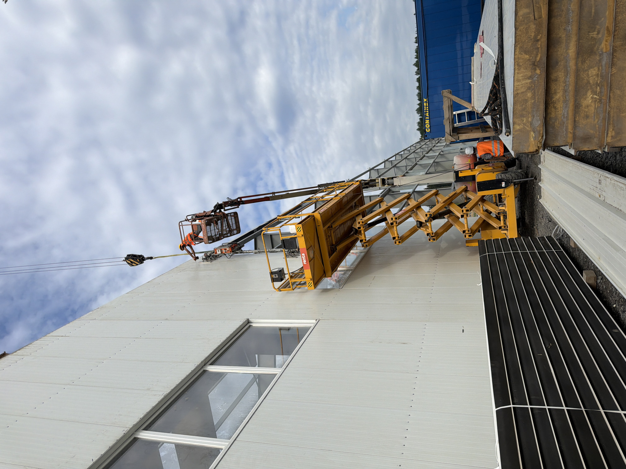 In Stróża, Poland, on a construction site, a scissor lift is positioned against the side of a large, modern building. The building features a light-colored, ribbed exterior wall and several large, rectangular windows. Two workers, wearing safety vests and hard hats, are operating the scissor lift. One worker is in the elevated platform of the lift, appearing to be working on the building's facade. The other worker is at the base of the lift, likely controlling its movement or assisting. Above the scissor lift, a crane hook is visible, suggesting ongoing material lifting operations. In the background, a large blue shipping container with the text "CONTAINEX" is parked, and stacks of building materials, including what appear to be insulated panels, are present on the ground. The sky is partly cloudy.