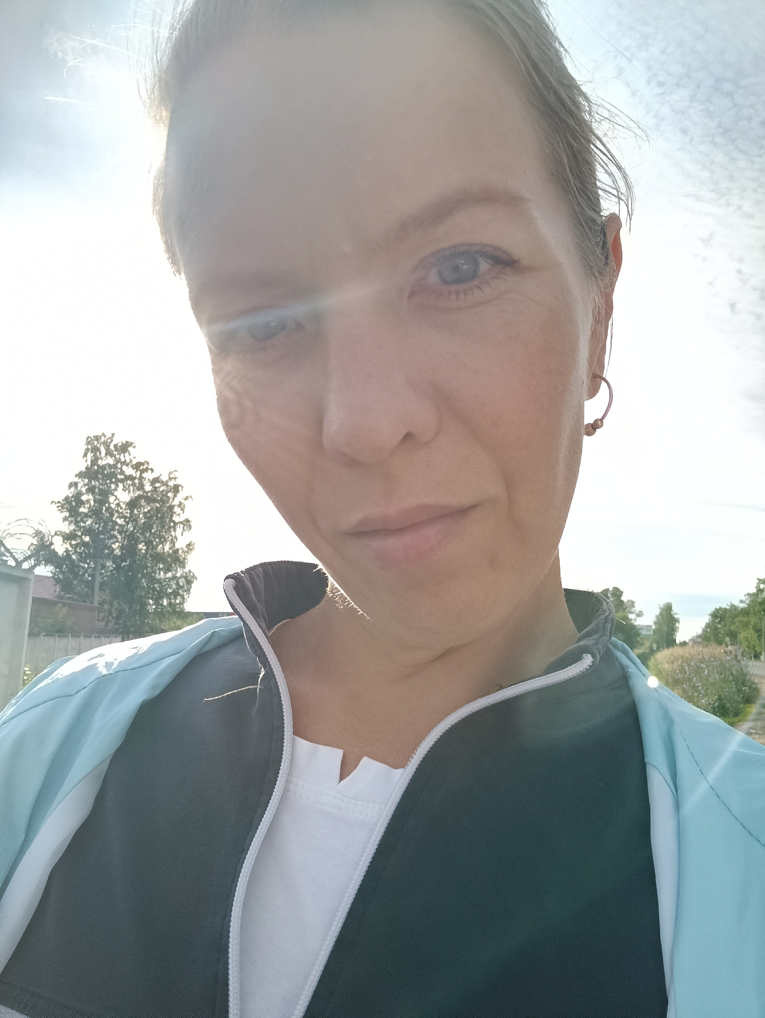 This is a close-up, low-angle selfie of a woman with fair skin and light brown hair, possibly taken outdoors during the daytime, as indicated by the bright sunlight and lens flare. She is wearing a light blue and dark gray zip-up jacket over a white t-shirt. She has light blue eyes and a single hoop earring in her right ear. The background is slightly blurred, showing some trees and vegetation, suggesting a natural or park-like setting. The woman's expression appears neutral or slightly contemplative. There is no visible text in the image. The sun is positioned to the left, casting a strong light and creating a flare across the top and left side of the image, obscuring some details of the sky and the woman's hair. The overall impression is of a casual outdoor moment, possibly during exercise or a walk.