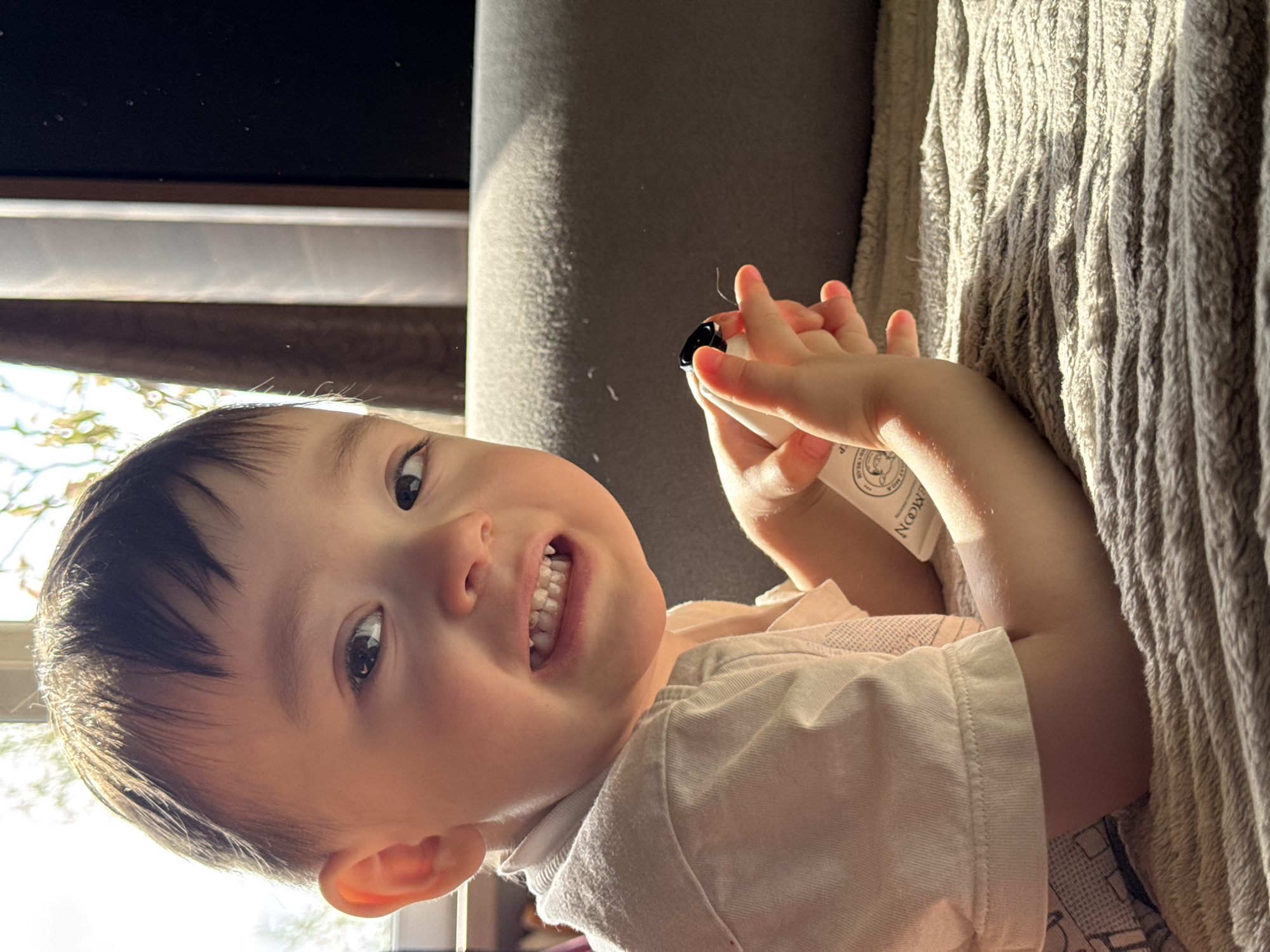 A young child lies on their back, holding a small white tube with a black cap. The child has dark hair and is wearing a light-colored shirt. The background includes a grey sofa and a textured blanket. The location in Izhevsk, Russia cannot be determined from the image.