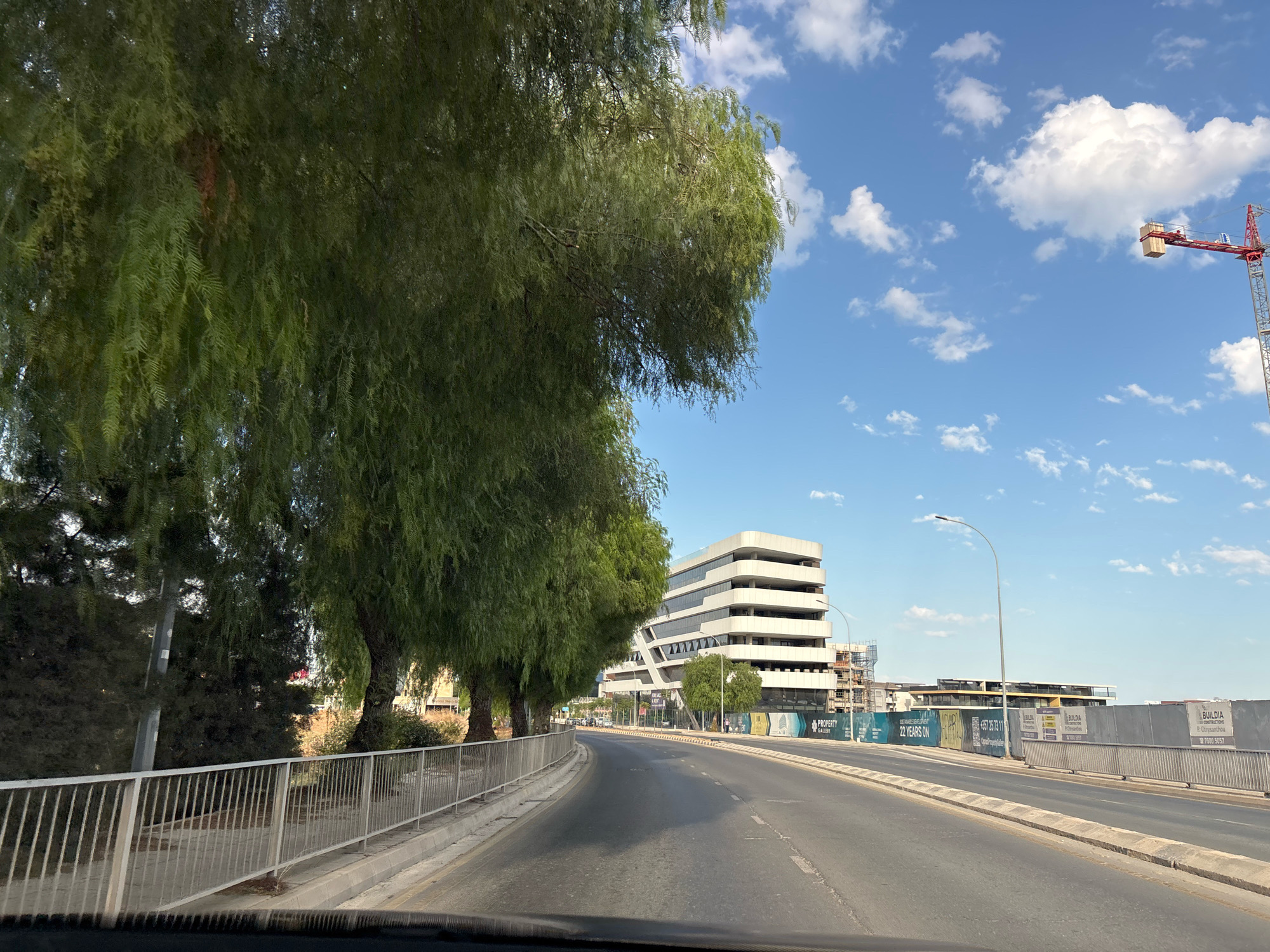 The image shows a road in Mesa Geitonia, Cyprus, with a large, modern white building on the right side of the road. The building has a distinctive, staggered design with multiple levels. In front of the building, there are construction barriers with advertisements and signs, indicating ongoing development. A red construction crane is visible in the distance above the buildings. On the left side of the road, there are several large trees with green foliage, casting shadows on the pavement. The sky is blue with scattered white clouds. The road itself appears to be a multi-lane street with white markings. A metal fence runs along the side of the road next to the trees. The overall scene suggests an urban environment with a mix of new construction and established greenery.