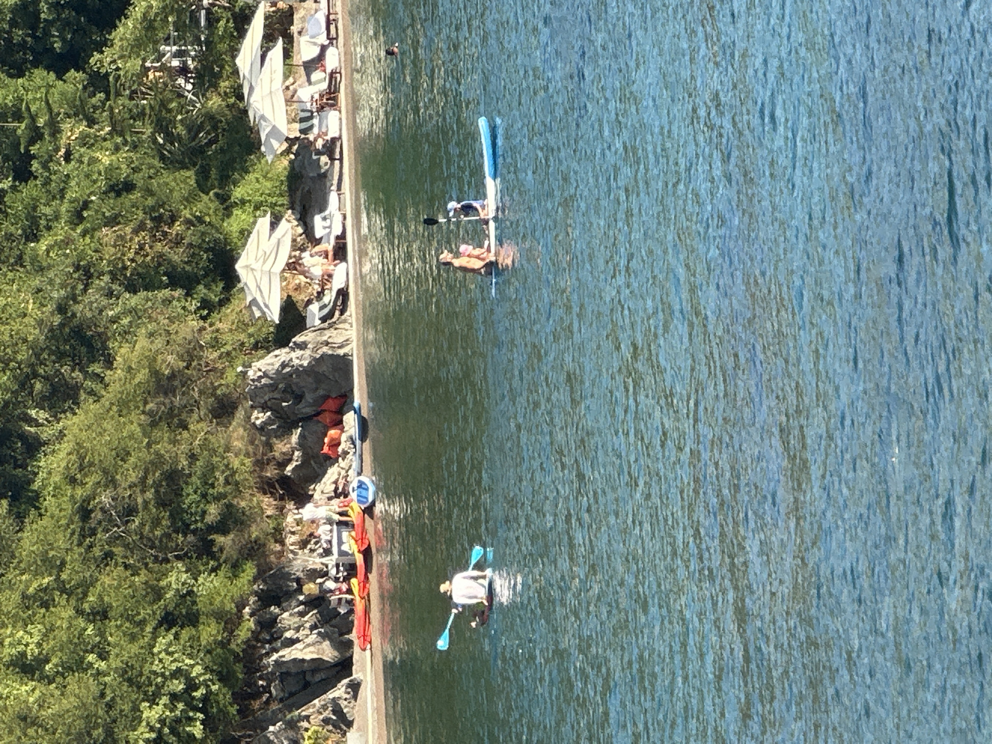 The image shows a beach scene in Budva, Montenegro. In the foreground, two people are paddleboarding on a calm, blue body of water. One person, wearing a light-colored shirt and a hat, is paddling a dark-colored paddleboard. The other paddleboard, which is light blue, has two people on it: an adult and a child. The adult is paddling with a black paddle. In the background, kayaks and other small boats are pulled up onto a sandy beach. Beyond the beach, there are rocks and lush green trees on a hillside. Several white umbrellas and lounge chairs are set up on the beach, suggesting a resort or leisure area. The overall scene depicts a sunny day with people enjoying recreational activities on the water and the beach.