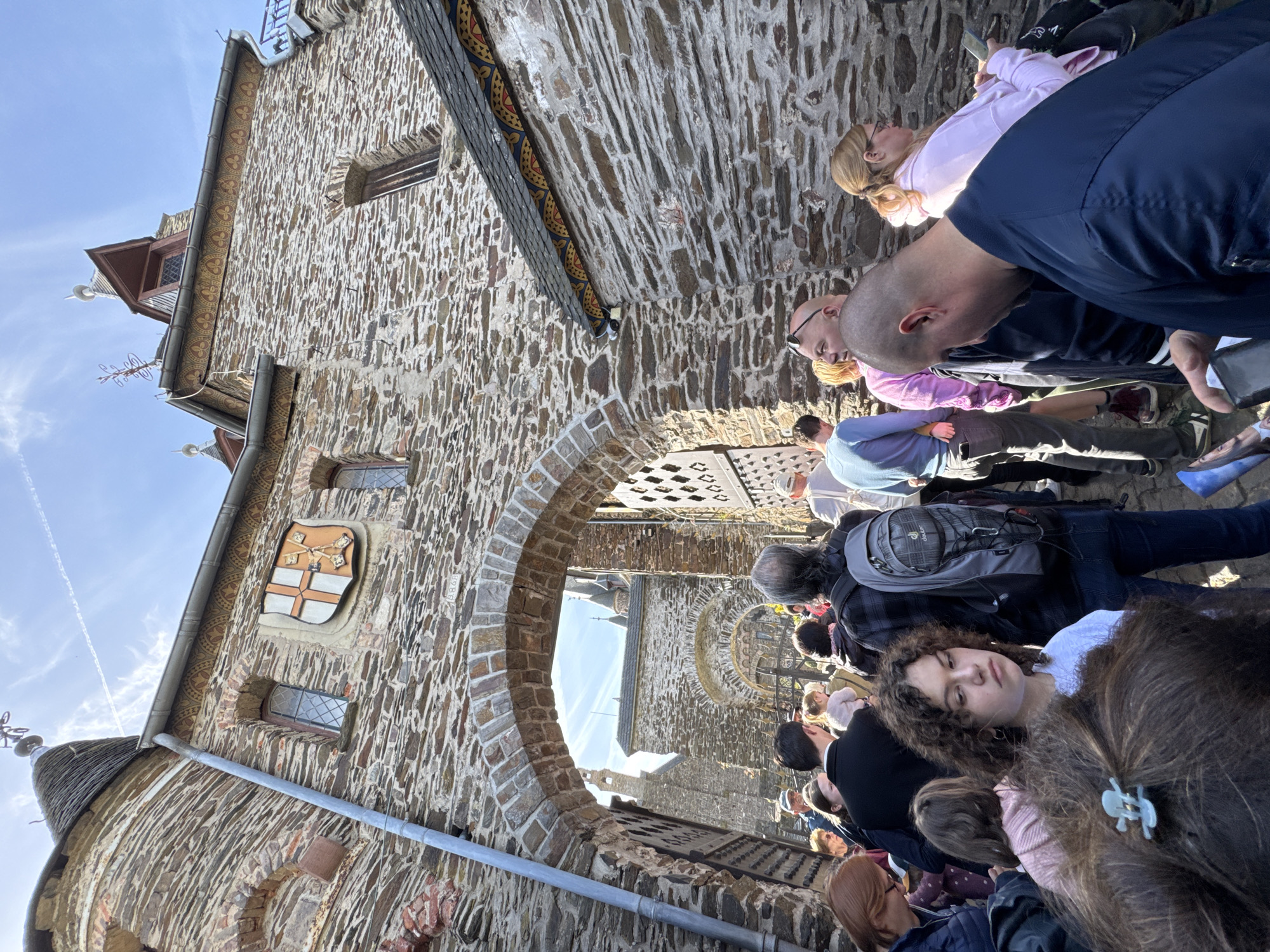 The image depicts a group of people standing in line outside the arched stone entrance to a building in Cochem, Germany. The building is constructed of rough-hewn stones, with a small window and a shield bearing a coat of arms visible on one side. The crowd appears to be made up of tourists or visitors waiting to enter the building, with people of various ages and clothing styles. Some are looking towards the building, while others are talking to each other. The sky is blue, suggesting it is a sunny day.