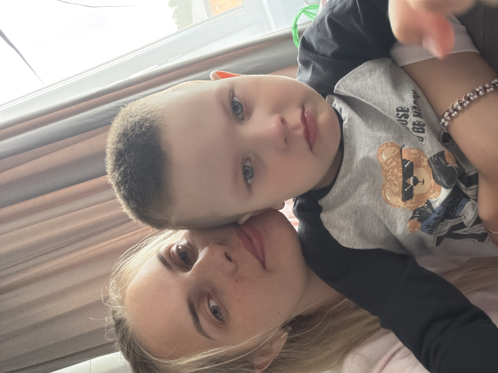 A woman with blonde hair is lying down with a young boy with short, light brown hair on top of her. The boy is wearing a grey t-shirt with a teddy bear graphic and black sleeves. The woman is wearing a bracelet on her right wrist. The location appears to be indoors, near a window with white frames. The photograph appears to be taken inside of an apartment or home. It is not possible to confirm that the location is in the city of Chaikovsky, Russia.