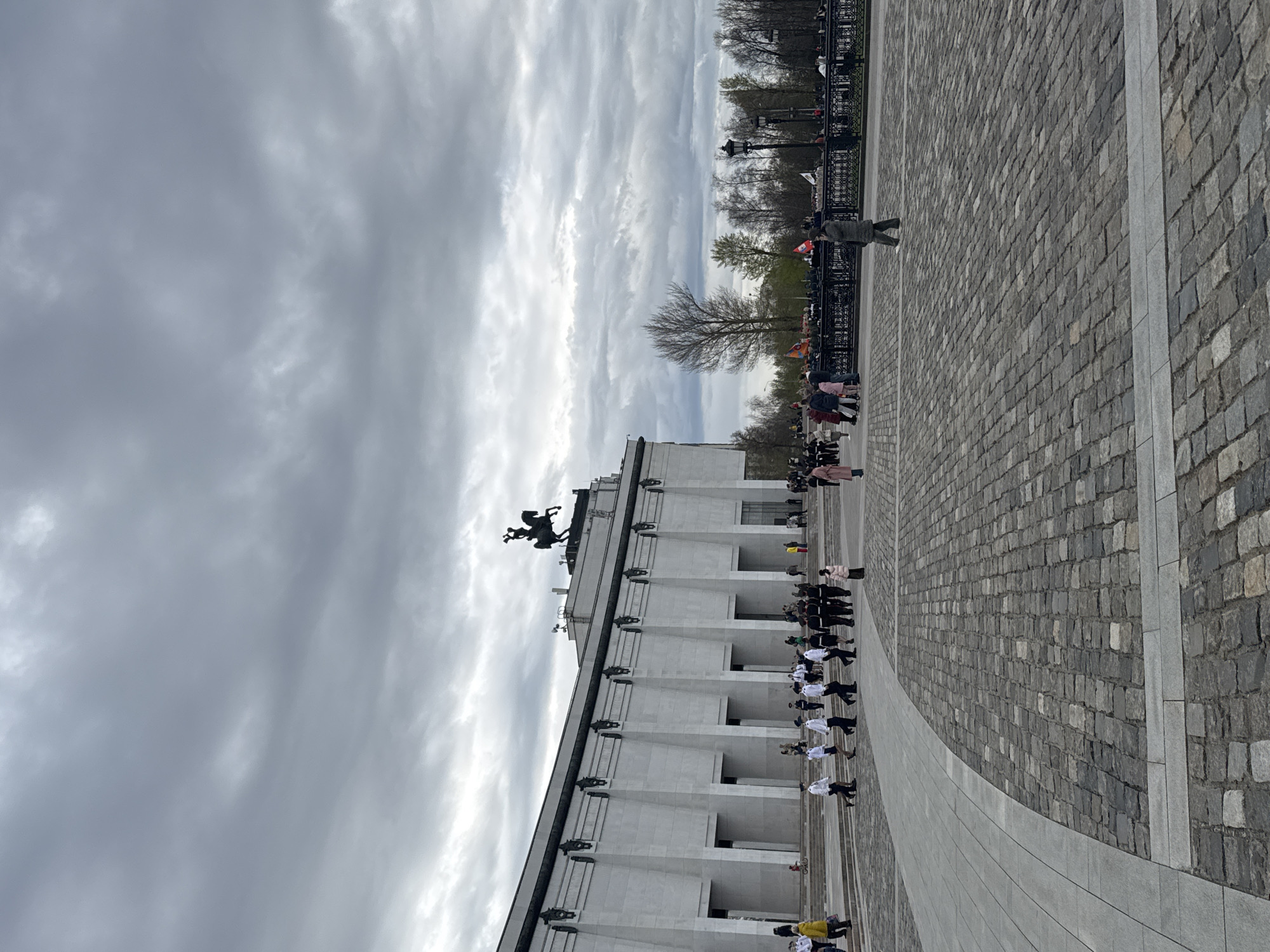 The image shows a view of Poklonnaya Hill in Moscow, Russia. A large, white monument stands on the left side of the image, topped with a statue of Saint George slaying a dragon. A line of people, some in white shirts and dark pants, stands in front of the monument. Other people are visible walking around in the background. The foreground is a paved area with a curved design. The sky is overcast with gray clouds. A row of trees and lampposts is visible in the distance.