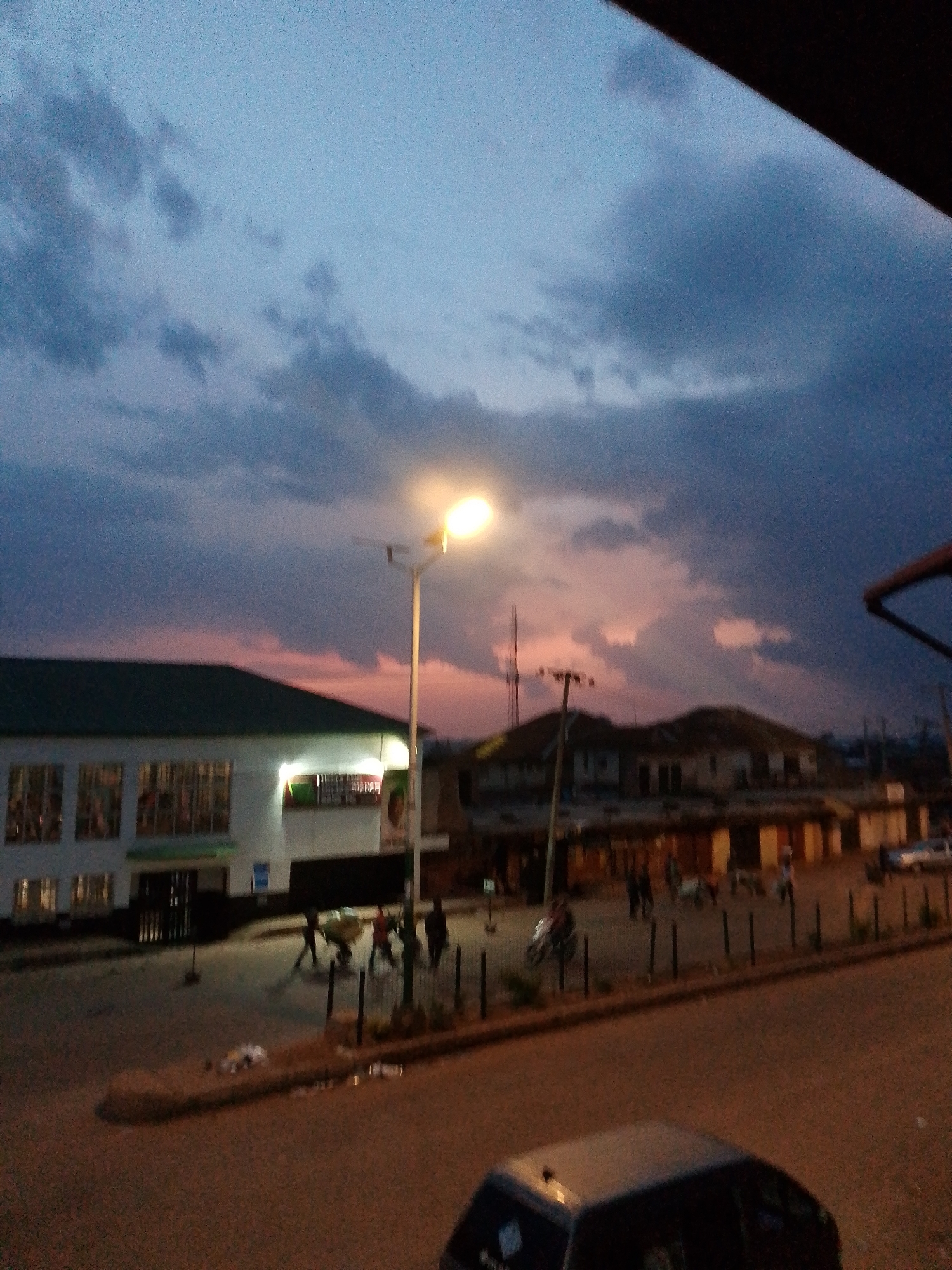 The image depicts a street scene in Bukuru, Nigeria, during dusk. A street light illuminates the foreground, casting a warm glow on the asphalt road. In the midground, a building with a green roof and large windows stands to the left, appearing to be a commercial establishment. Several people are visible on the street, some appearing to be walking or engaged in activities. To the right of the building, more structures and a distant antenna are visible. The sky above is filled with dramatic clouds, painted with hues of purple, pink, and blue, indicative of the sunset. In the lower right corner of the image, a car is parked. The overall impression is of a typical urban evening in Bukuru.