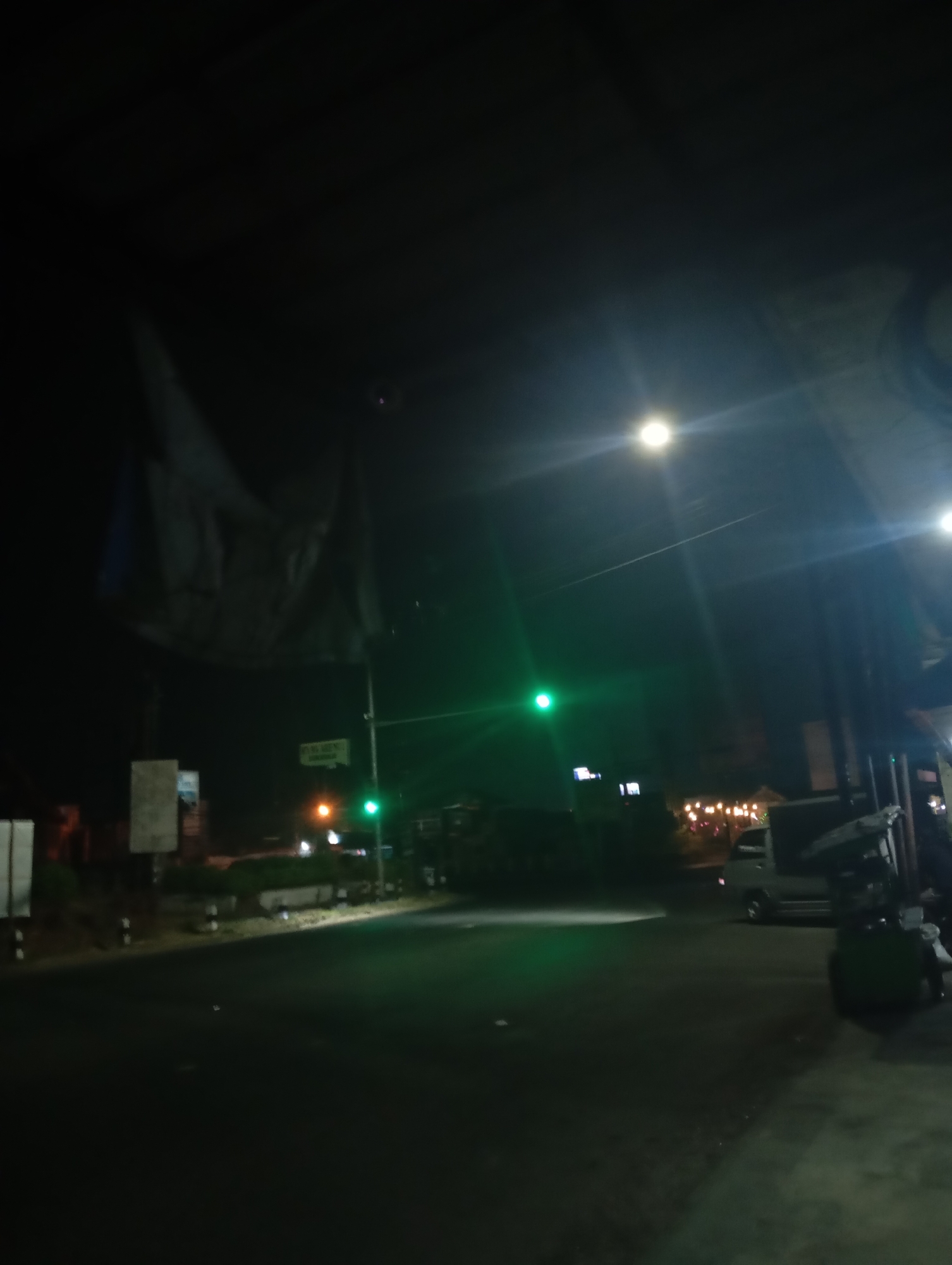 The image depicts a street scene at night in Sokaraja, Indonesia. A traffic light is visible, illuminated green, indicating that traffic can proceed. In the foreground, the dark asphalt road stretches into the distance. On the right side of the road, a portion of a building or structure is visible, along with a parked truck or van. A large, light-colored tarp or banner hangs from a pole on the left side of the street, obscuring part of the background. Several streetlights are also present, casting a glow on the scene and creating lens flares in the dark sky. The overall atmosphere is that of a quiet, dimly lit street in a town or city at night.
