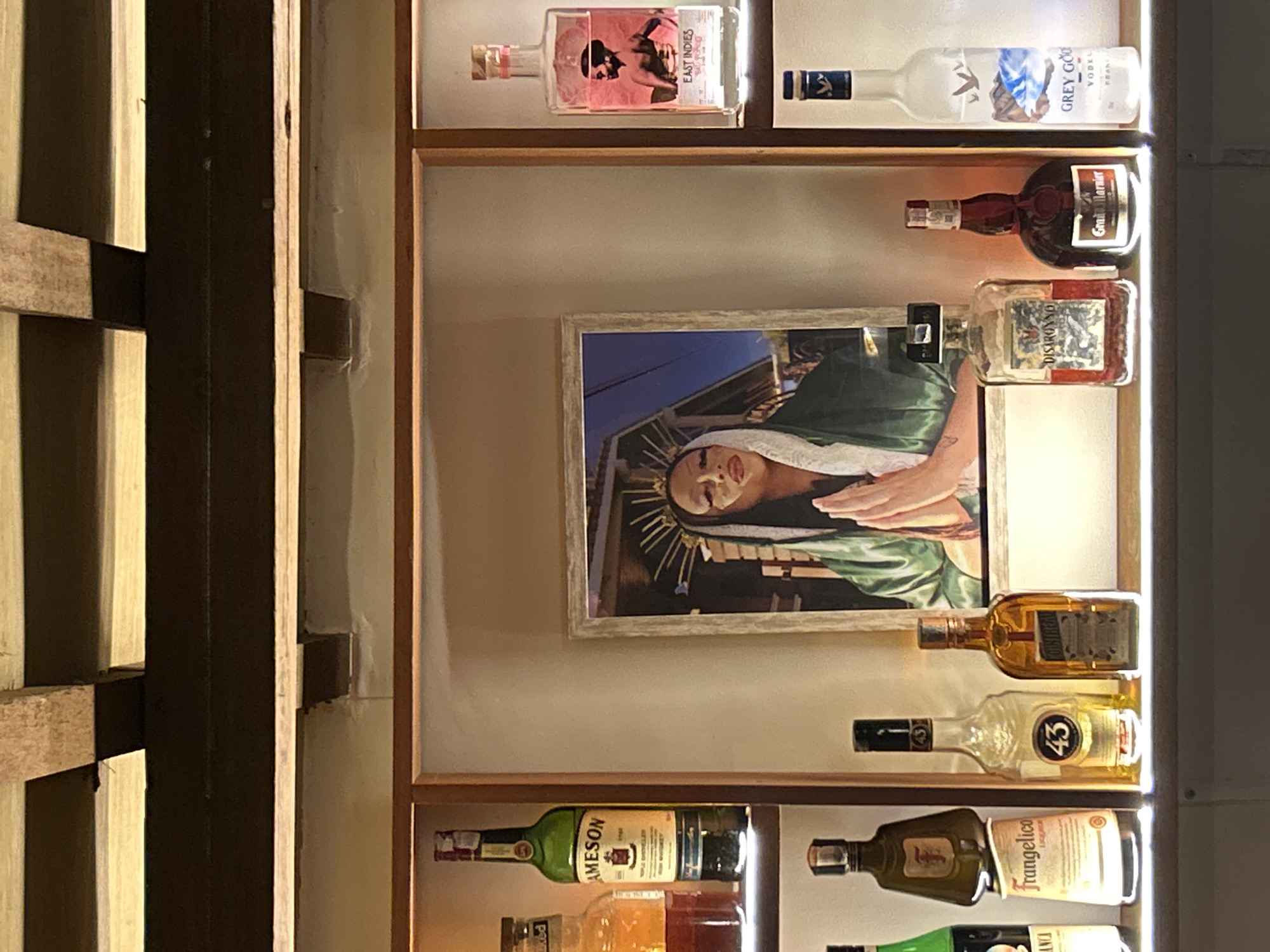 This is a medium shot, eye-level, indoor photograph taken in Kuta Selatan, Indonesia. The scene is a dimly lit bar or display shelf featuring various bottles of liquor and a framed picture.
Dominating the center of the frame is a framed picture depicting a person dressed in a green robe and white veil, with hands clasped in prayer. The person has striking makeup and a crown-like headpiece. The background of the picture appears to be an outdoor setting with a twilight sky.
Surrounding the central picture are several bottles of alcohol arranged on shelves. On the left shelf, from top to bottom, are a green bottle of Jameson Irish Whiskey, a clear bottle with amber liquid, and a dark bottle with a green label. To the right of the Jameson bottle is a bottle of Frangelico. Further right are two clear bottles, one labeled "43" and another with an amber liquid labeled "Cointreau".
On the right shelf, from top to bottom, is a tall, clear bottle with a pink label featuring a silhouette of a person, labeled "EAST INDIES RUM". Below that is a clear bottle with a label depicting mountains and birds, labeled "GREY GOOSE VODKA". Between the picture and the Grey Goose bottle, there are two smaller bottles, one labeled "DISARONNO" and another dark bottle labeled "Grand Marnier".
The shelves appear to be made of wood, and the overall lighting suggests an evening or interior setting designed for ambiance. The background above the shelves shows wooden beams and a dark ceiling.