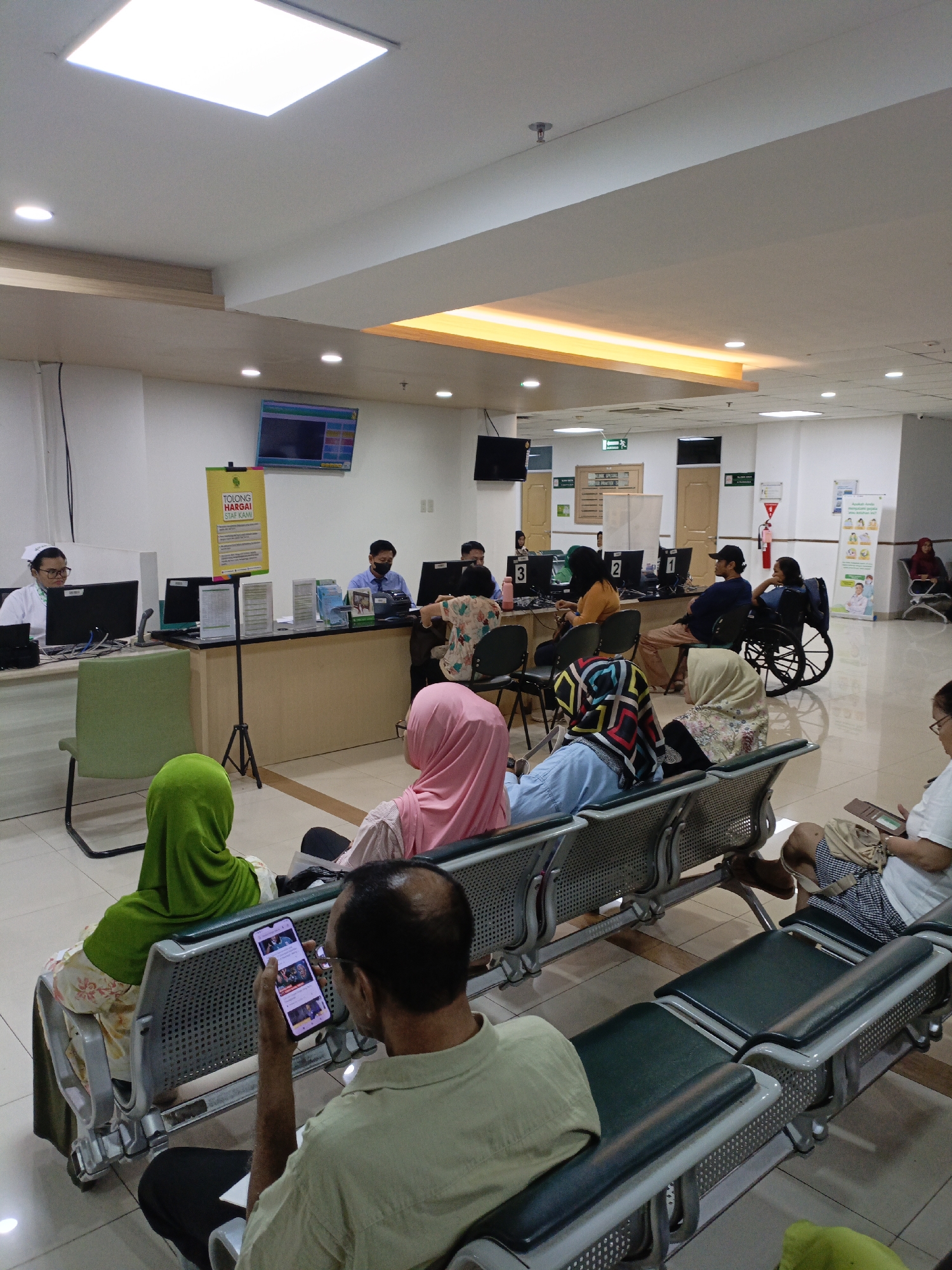 The image depicts a waiting area in what appears to be a clinic or hospital in East Jakarta, Indonesia. Several people are seated on metallic benches, some facing a counter where staff are attending to them. A nurse in uniform is visible behind a computer terminal on the left. A man in the foreground is looking at his mobile phone. Another individual is seated in a wheelchair. The environment is brightly lit with fluorescent ceiling lights and features a reception desk with multiple computer monitors and information posters. The overall scene suggests a typical day at a healthcare facility's waiting room.