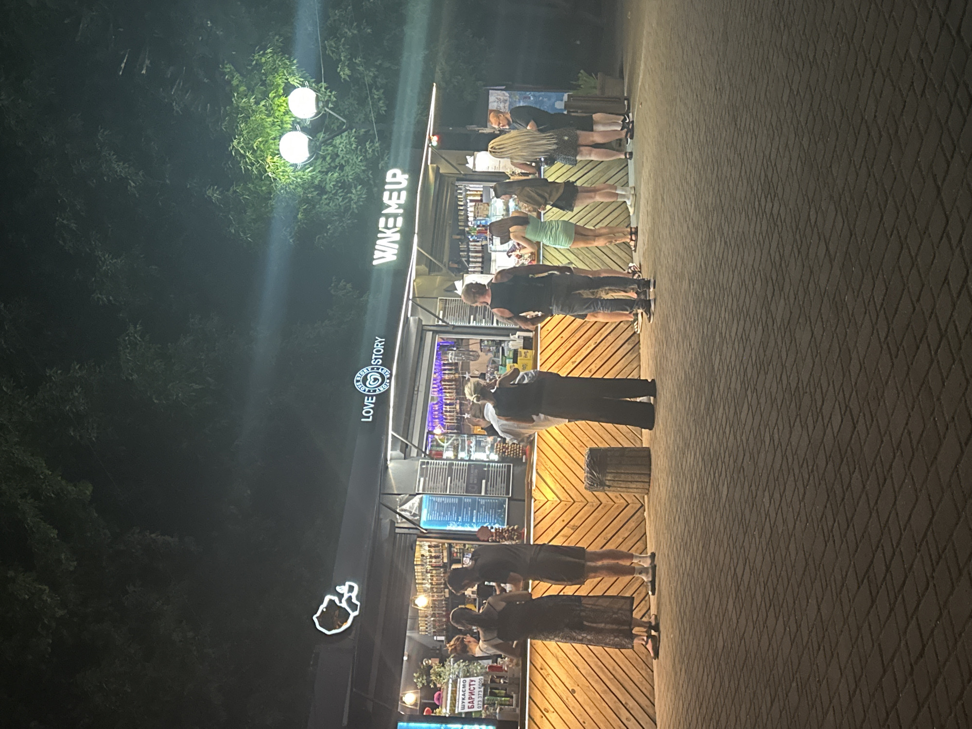 This is a nighttime outdoor shot of a modern outdoor cafe or kiosk with illuminated signage in Odesa, Ukraine. The cafe features a wooden facade with a chevron pattern and has a long counter where several people are standing, appearing to order or receive items. There are multiple individuals in the scene, some standing in line, and others in conversation. The signage includes "LOVE STORY" and "WAKE ME UP." The environment is dark with artificial lighting illuminating the cafe and the people. The ground is paved with interlocking stones. The scene appears to be a public space, possibly a promenade or pedestrian area, with trees visible in the background.
