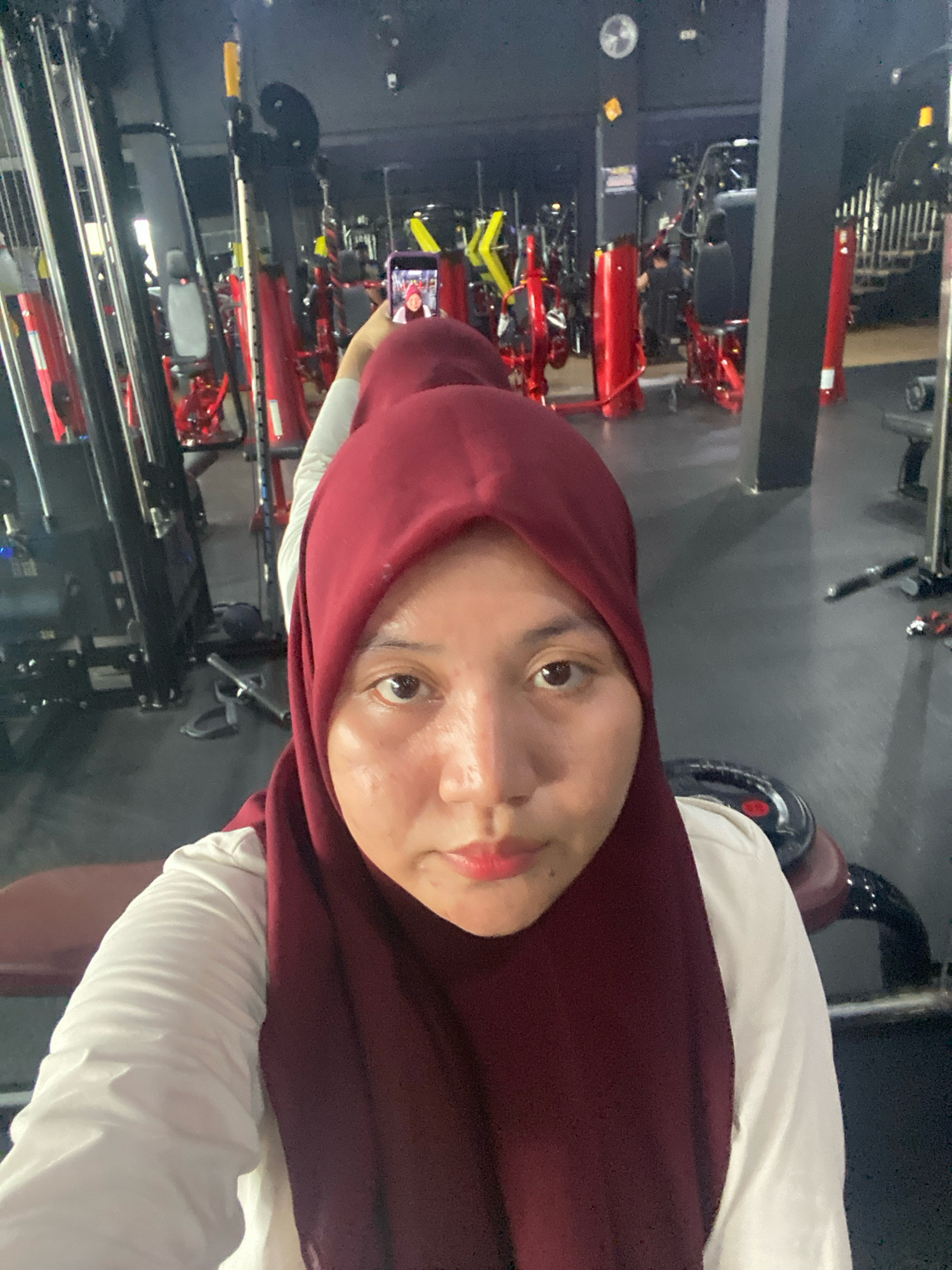 A young woman wearing a maroon hijab and a white shirt is taking a selfie in a gym. She is holding a smartphone in her right hand. The gym is equipped with various weight machines, all in red and black. The floor is dark and reflective, suggesting it is polished concrete or a similar material. The lighting is artificial, typical of an indoor gym environment. The woman's facial features are visible, and she appears to be looking at the phone's screen. In the background, other gym equipment and possibly other patrons can be seen, though they are out of focus. The scene captures a moment of personal activity within a public fitness facility. The location is identified as Banjarbaru, Indonesia.