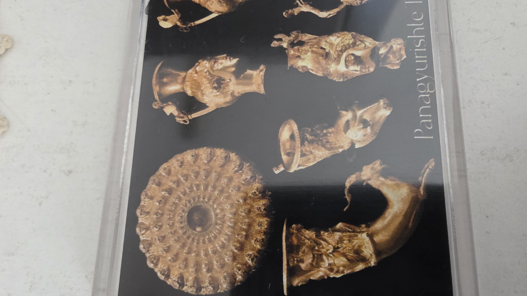 The image shows a photograph of the Panagyurishte Treasure, a Thracian gold hoard, encased in a plastic display. The objects depicted include a rhyton in the shape of a ram's head, a rhyton in the shape of a deer's head, a vessel or amphora, and a phiale (shallow dish) adorned with concentric rows of embossed acorns. The inscription "Panagyurishte" is visible. The objects appear to be displayed in a museum. The location is likely the Regional Historical Museum of Blagoevgrad, Bulgaria, which houses a replica of the treasure.