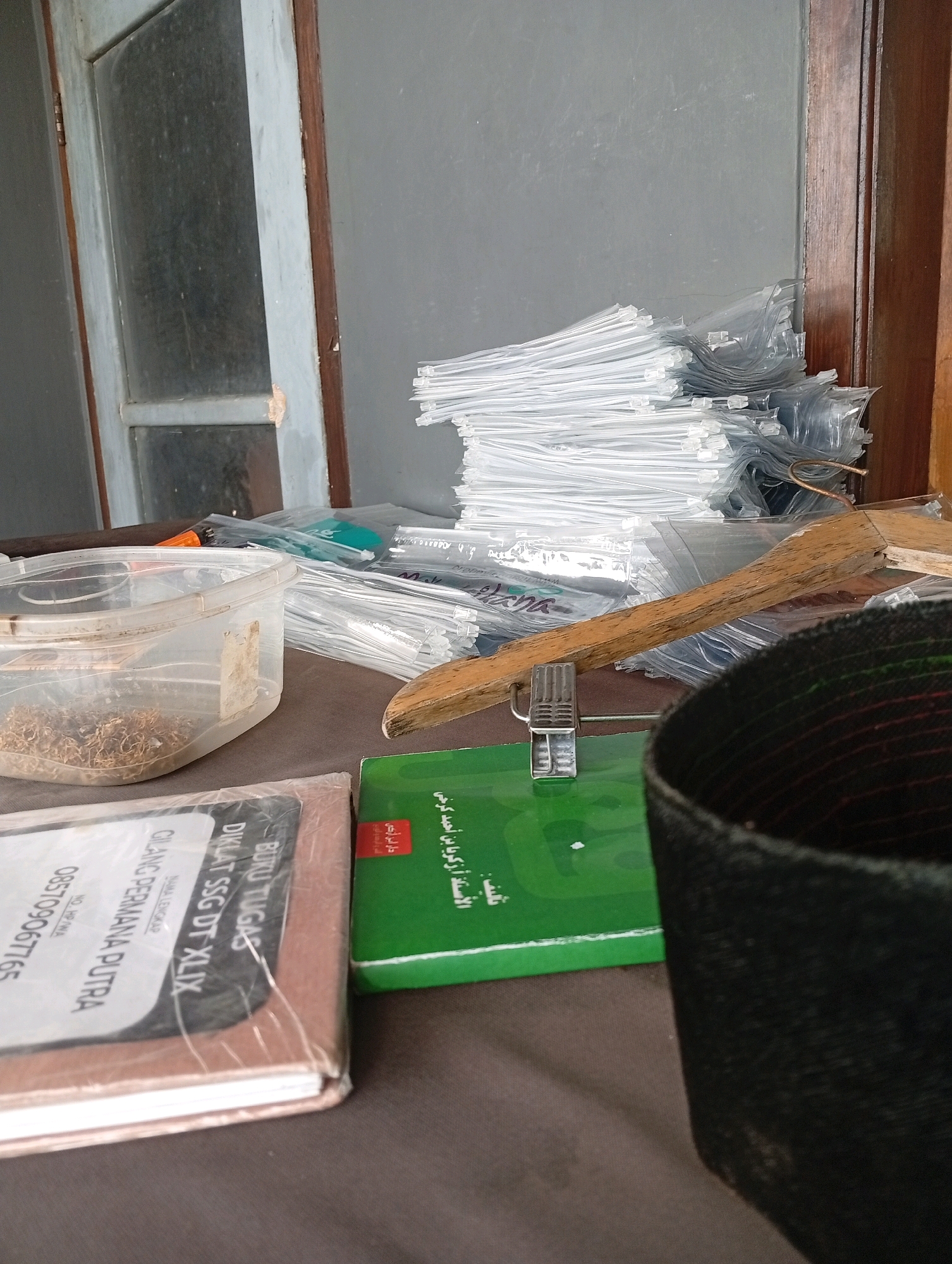 The image depicts a cluttered surface, likely a table, with various items. In the foreground, a clear plastic container holds dried tobacco leaves. To the left, a book with a brown cover is visible, featuring text in white and black, including what appears to be a phone number and a name. A green book with Arabic text is placed next to it. Behind these items, a large stack of transparent plastic bags, possibly for packaging, is piled high. A wooden hanger rests horizontally across some of the bags. In the upper left portion of the image, a partially visible window with a light blue frame and wooden trim is seen. The background is a plain, dark grey wall. The overall scene suggests a workspace or storage area, possibly in a home or small business. The specific location in Tasikmalaya, Indonesia, is not discernible from the visual information provided.
