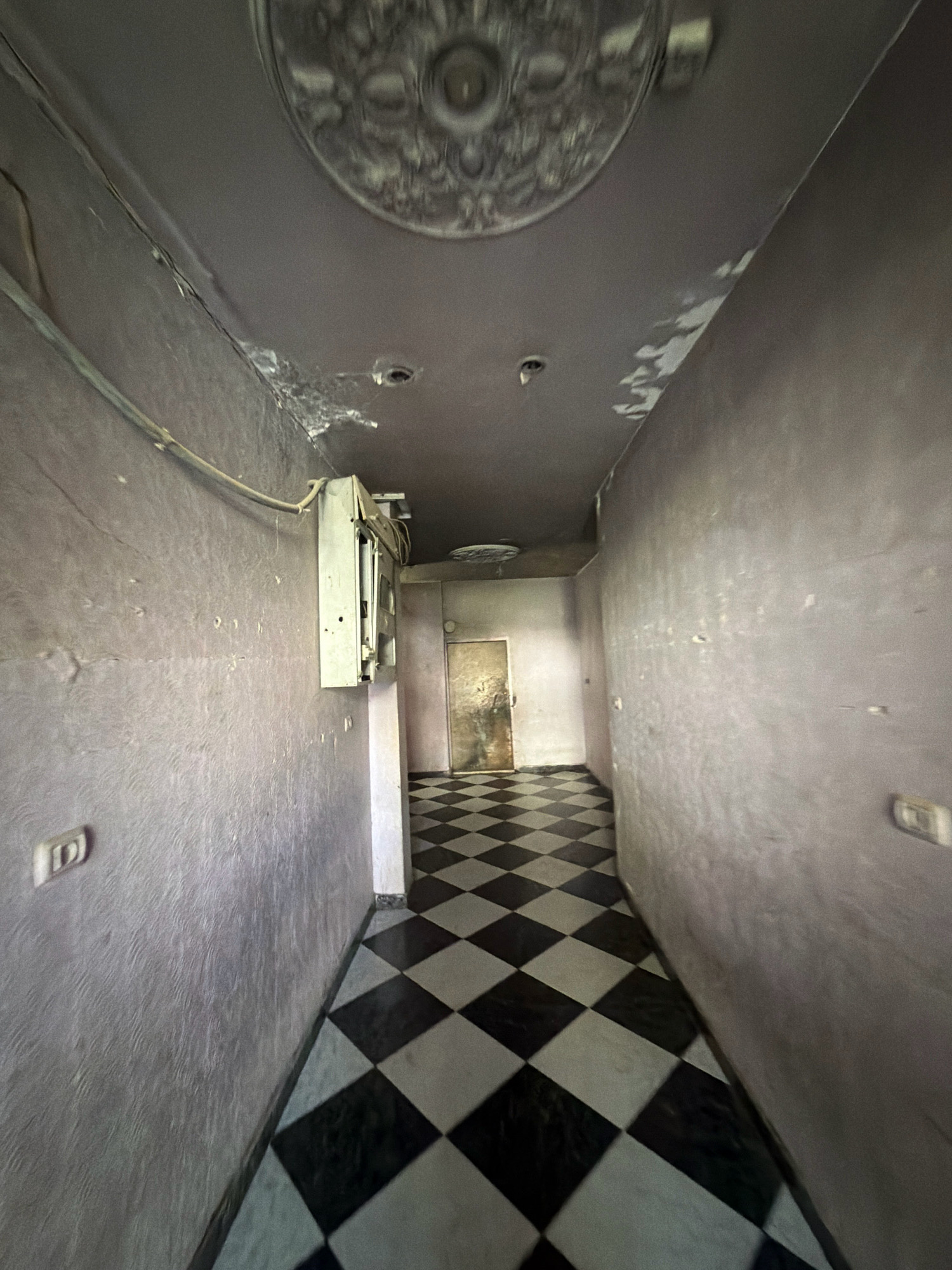The image shows an interior hallway in a building in Gaza, Palestinian Territories. The hallway has a checkerboard floor with black and white tiles. The walls are painted a pale purple color and appear to be textured. There is a light fixture on the ceiling, and what appears to be an air conditioning unit mounted on the wall. A closed door is visible at the end of the hallway. The scene appears to be in a state of disrepair, with peeling paint and visible cracks in the walls. The lighting is dim, suggesting it may be day or evening.