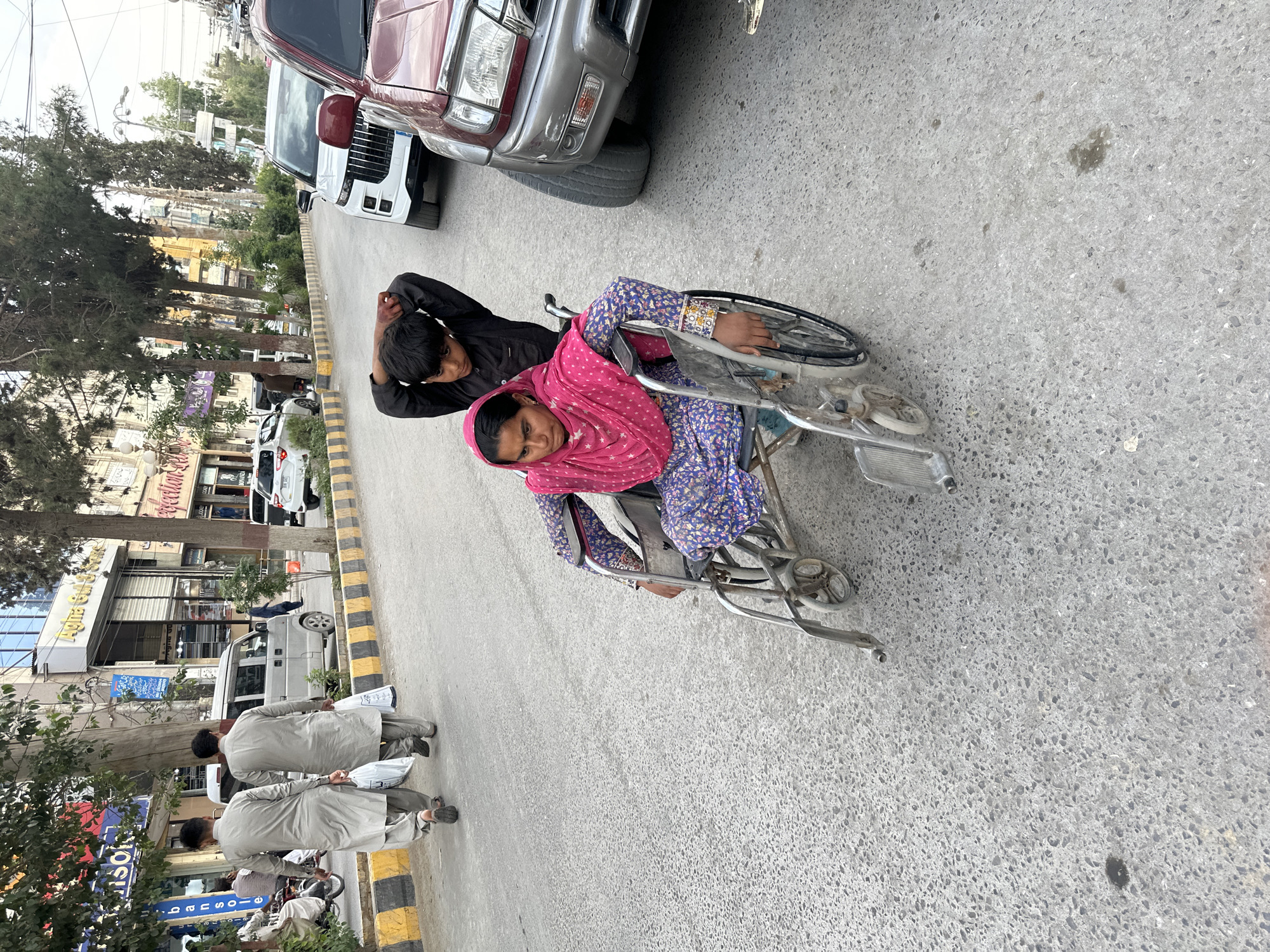 The image shows a woman in a wheelchair on a street in Quetta City Tehsil, Pakistan. She wears a pink headscarf and a blue floral patterned dress. A boy in a black shirt is leaning on the back of her wheelchair. They are on an asphalt road next to a sidewalk with yellow and black striped paint. Cars are parked on the side of the road, and buildings with shops and businesses are visible in the background. Several men in light-colored traditional shalwar kameez are walking on the sidewalk.