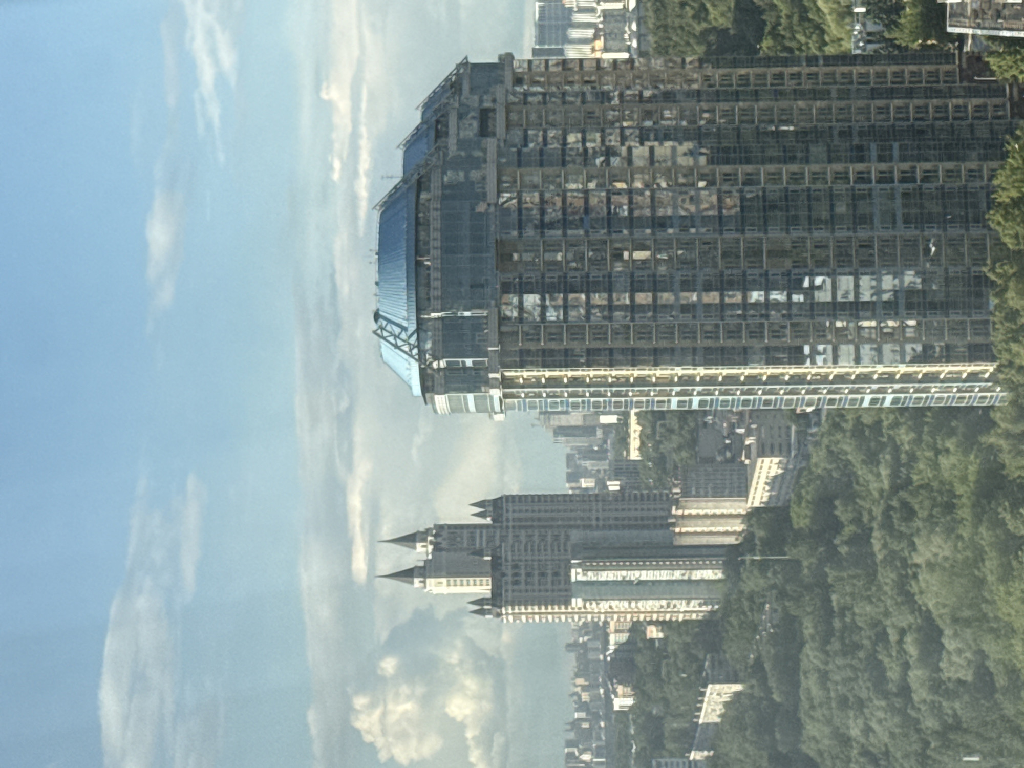 The image shows a view of Khimki, Russia. In the foreground, a modern, glass-faced skyscraper dominates the skyline. Adjacent to it, another large, older building with architectural details and spired roofs is visible. The cityscape is surrounded by lush green trees. The sky above is light blue with scattered clouds.