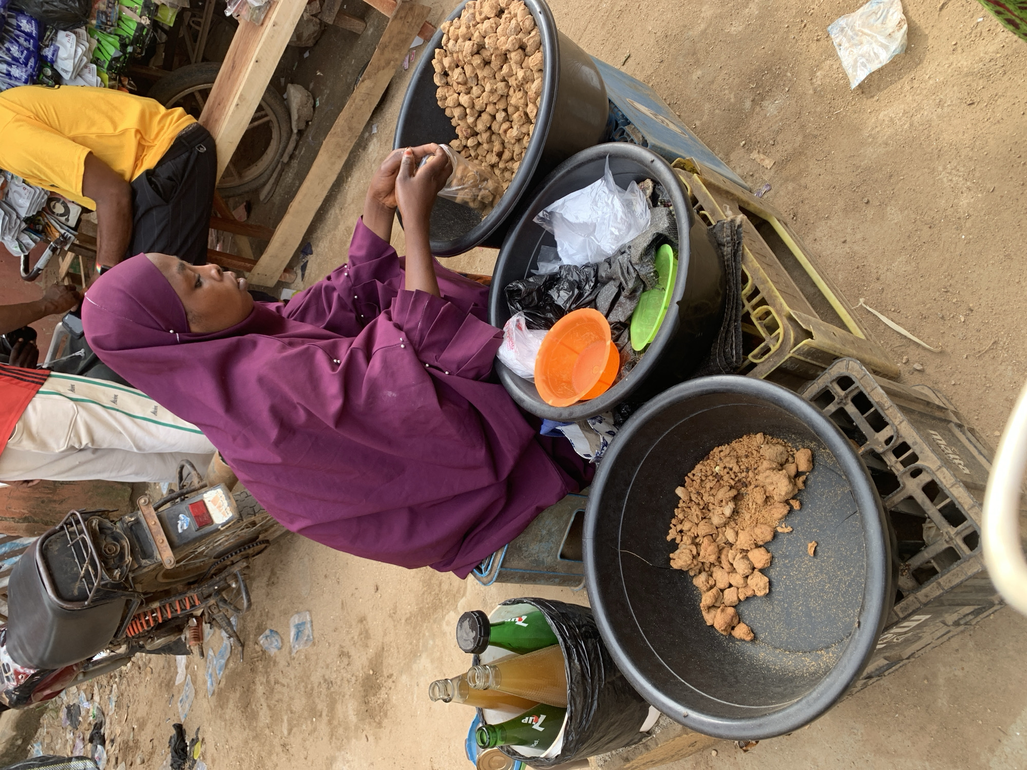 The image depicts a street scene in what appears to be a marketplace in Kafanchan B, Nigeria. A woman wearing a purple hijab is seated beside three large black plastic tubs, filled with what appear to be edible goods. One tub contains tan, roughly spherical objects, possibly fried dough or nuts. A second tub holds a mix of items, including orange and green plastic containers, along with clear and black plastic bags. The third tub contains a brown, lumpy substance, possibly a type of bean or seed. The woman appears to be handling the food, possibly portioning it for sale. Beside the tubs is a stack of empty plastic crates and a black plastic bag holding several glass bottles, possibly soft drinks or other beverages. A man in a yellow shirt is partially visible in the upper left, sitting on a low wooden structure. In the lower left, part of a motorbike is visible, suggesting transport to and from the market. The ground is dirt and scattered with debris.